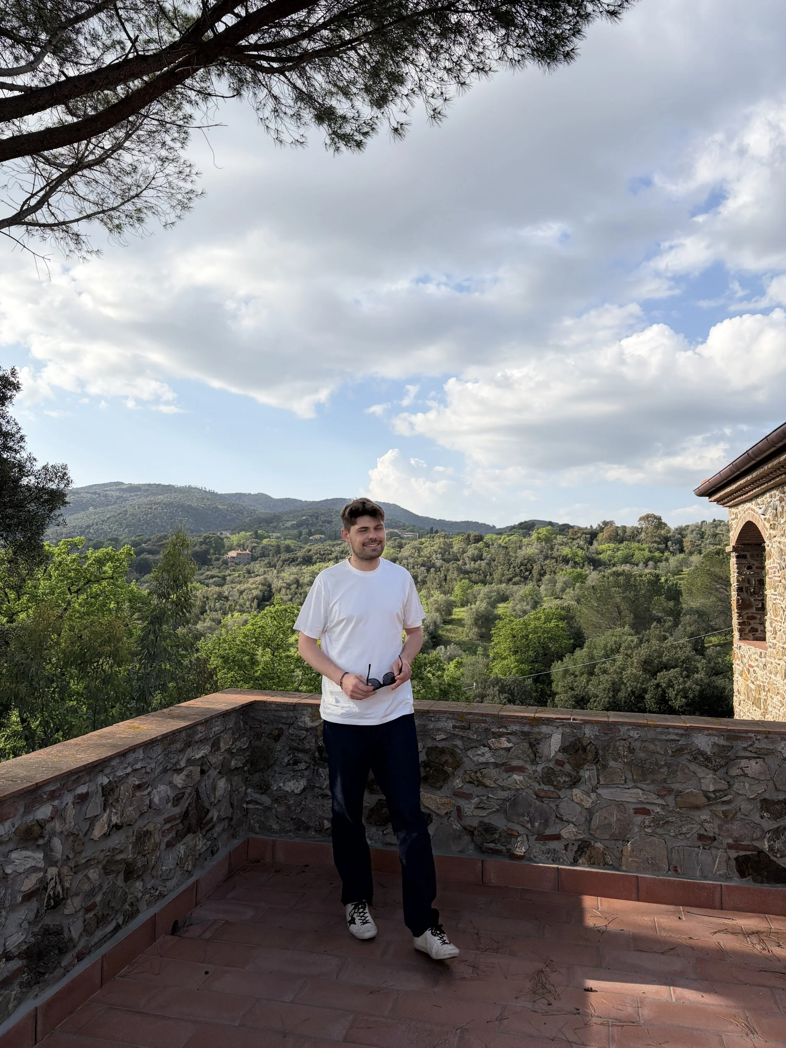 Smiling man in white t-shirt and dark pants standing on a stone balcony with a scenic view of rolling hills and blue sky with scattered clouds, holding black sunglasses.