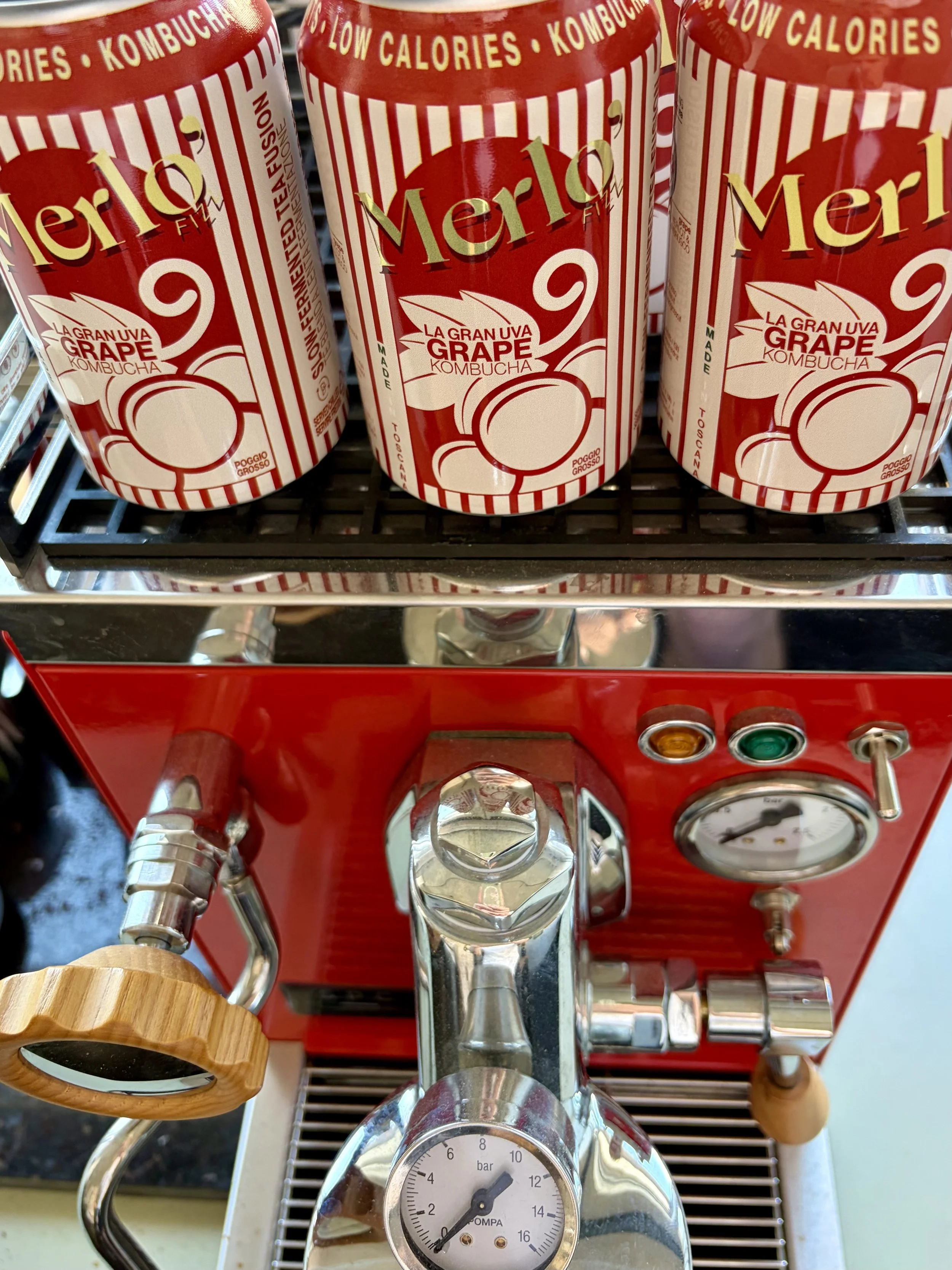 Red beverage dispenser with a wooden handle, labeled 'Merlo' and 'Grape Kombucha,' sitting on a metal stand with a pressure gauge reading around 6 bar, surrounded by metal pipes and a red control panel with gauges and switches.