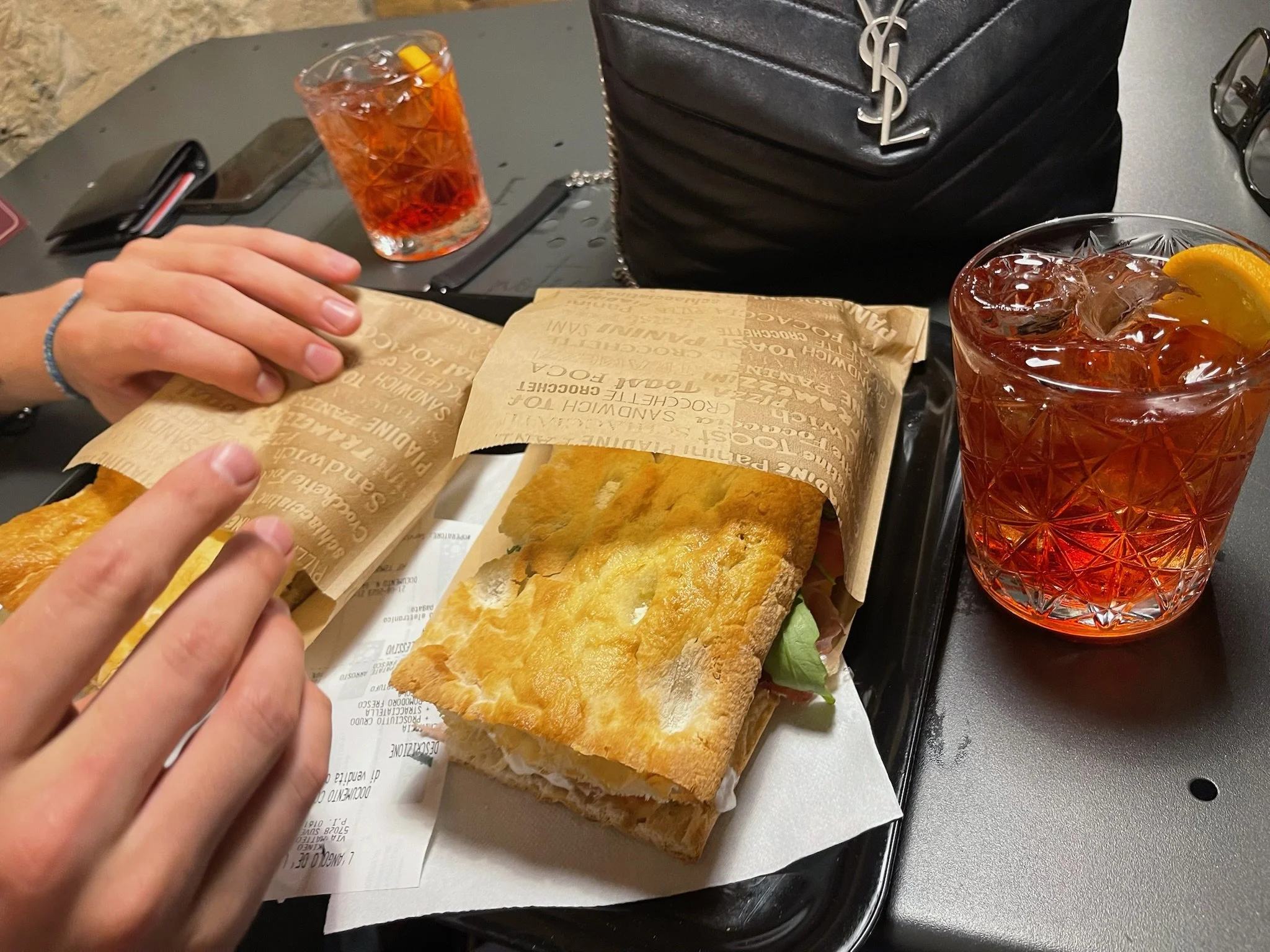 A tray with a toasted sandwich or panini and a sandwich wrap, two glasses of iced drinks with orange slices, and a black purse on a dark table.
