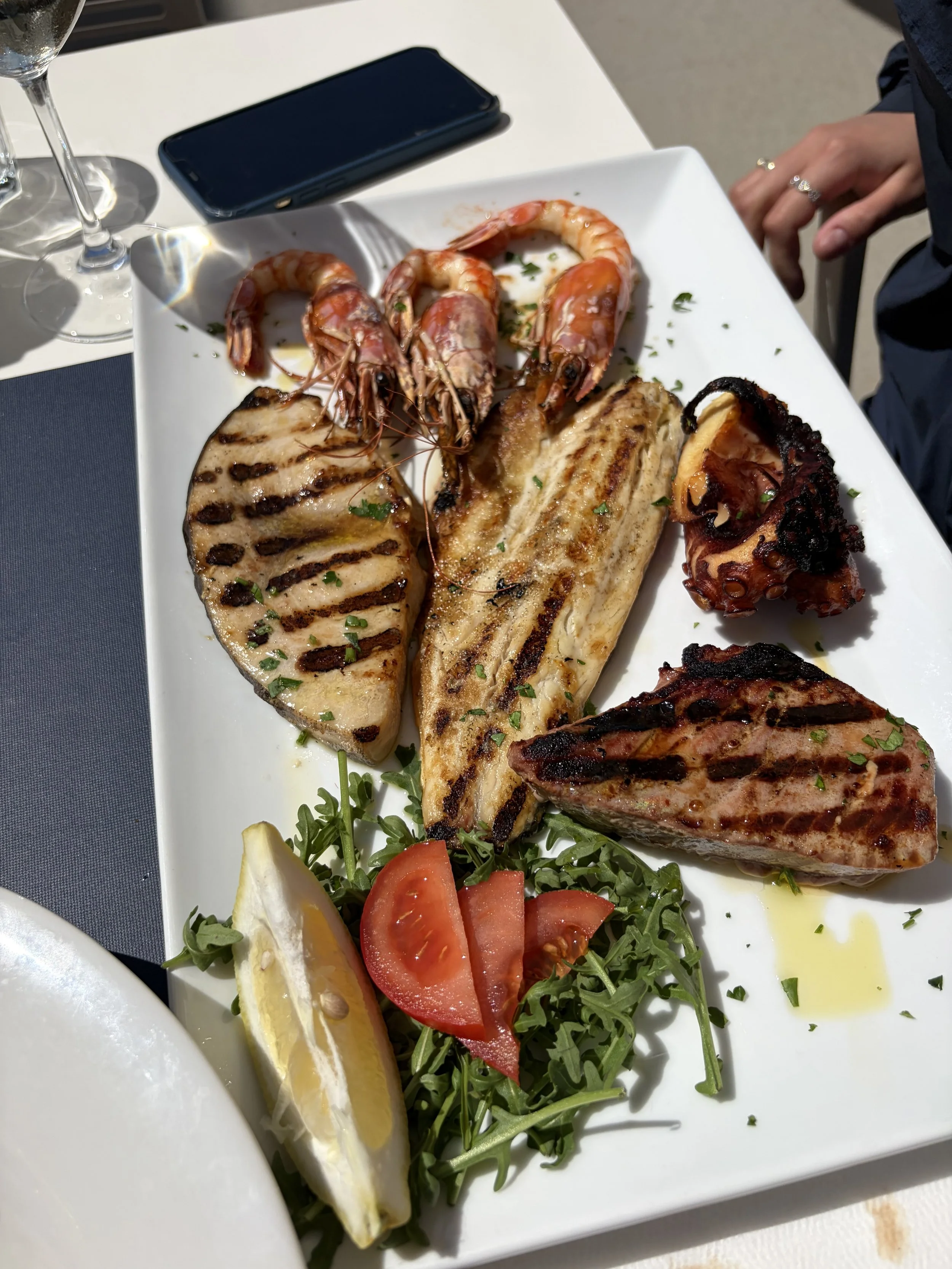 A white rectangular plate with grilled fish, grilled shrimp, and grilled octopus, garnished with fresh parsley, with lemon wedges, cherry tomato slices, and arugula salad on the side.