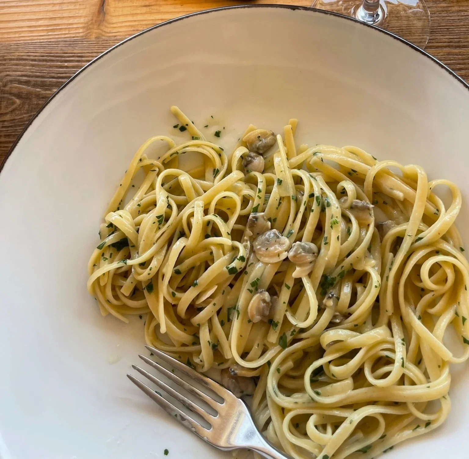 Plate of spaghetti pasta with clams and chopped herbs, served with a fork and a glass of wine in the background.