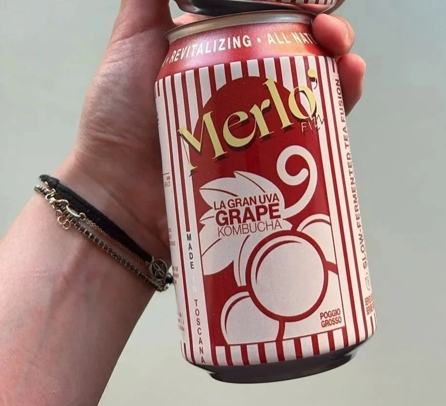 A hand holding a can of Merlot grape kombucha with a white and red striped design and text.