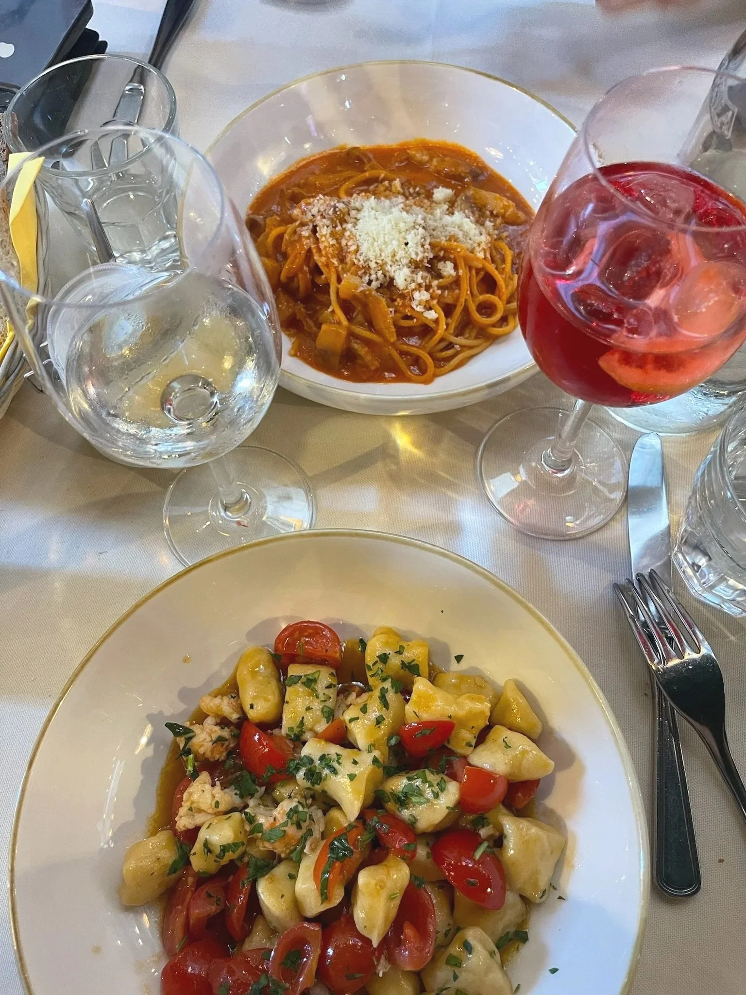 A table with a plate of cherry tomato and pasta salad, a bowl of spaghetti with tomato sauce topped with cheese, and a glass of red drink with ice, surrounded by empty glasses and silverware.