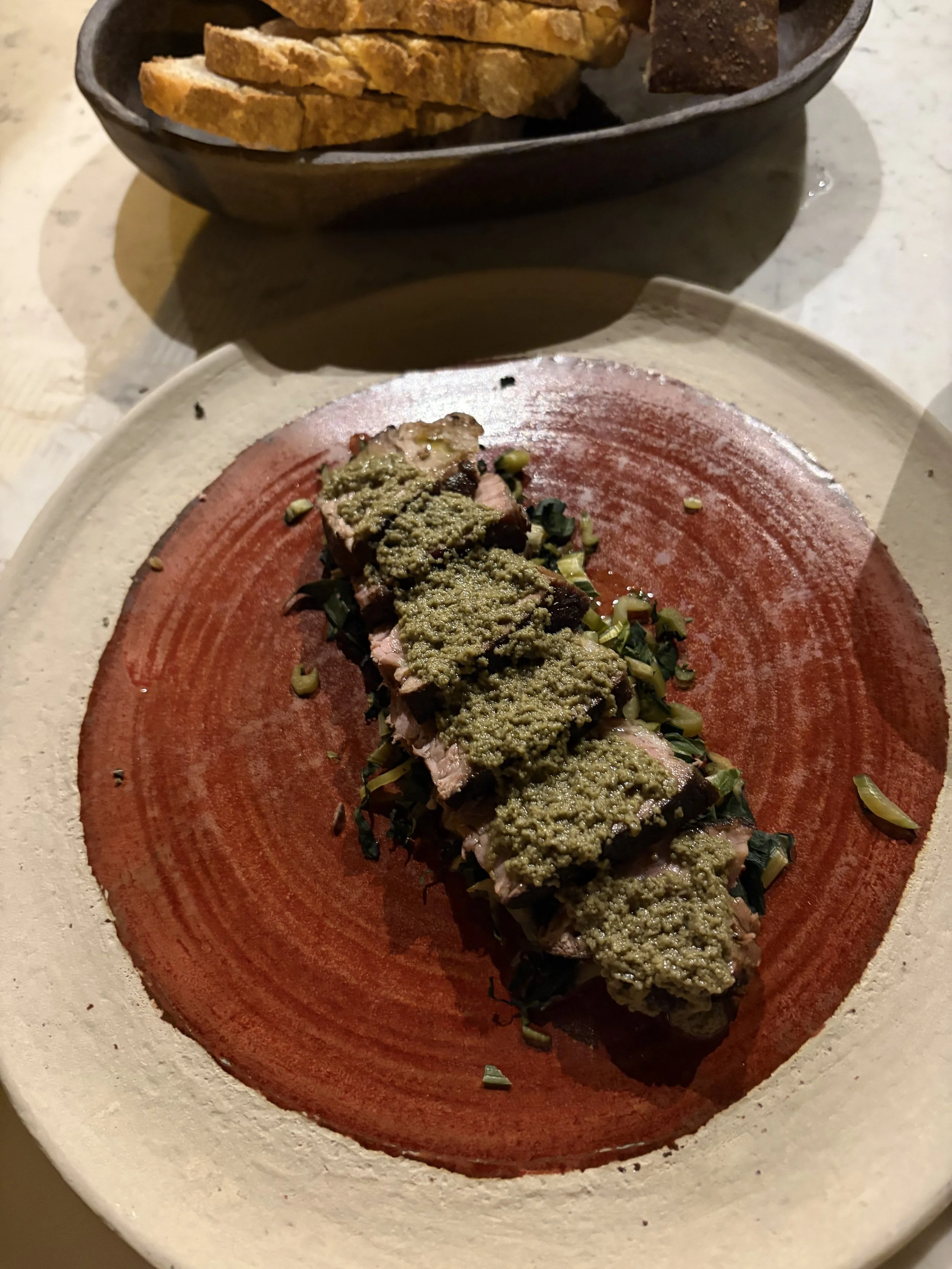 A plate with sliced meat topped with green sauce and chopped herbs, in front of a black bowl with sliced bread.