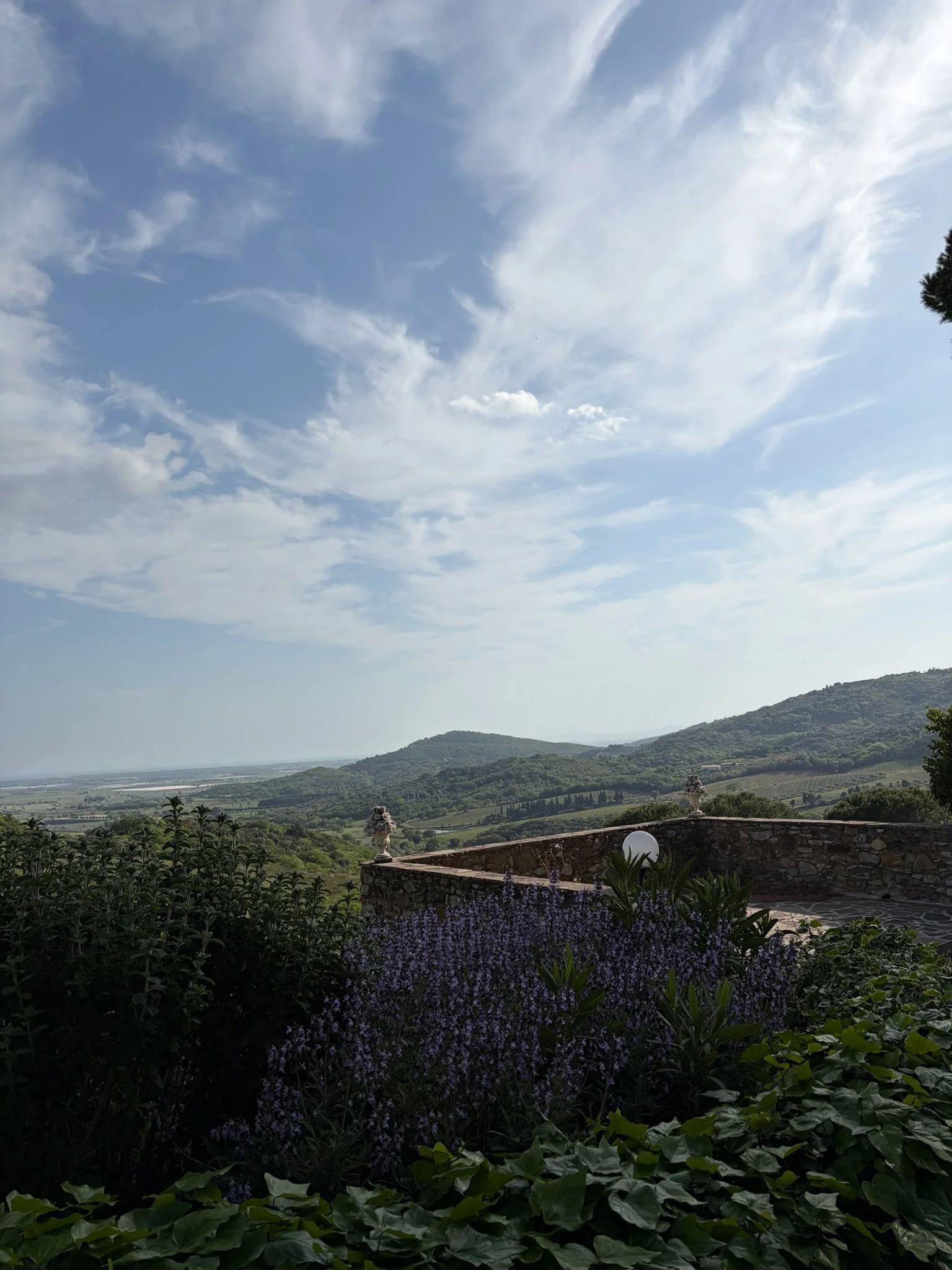View of a hilly landscape under partly cloudy sky, with a stone wall, decorative urns, and purple flowers in the foreground.