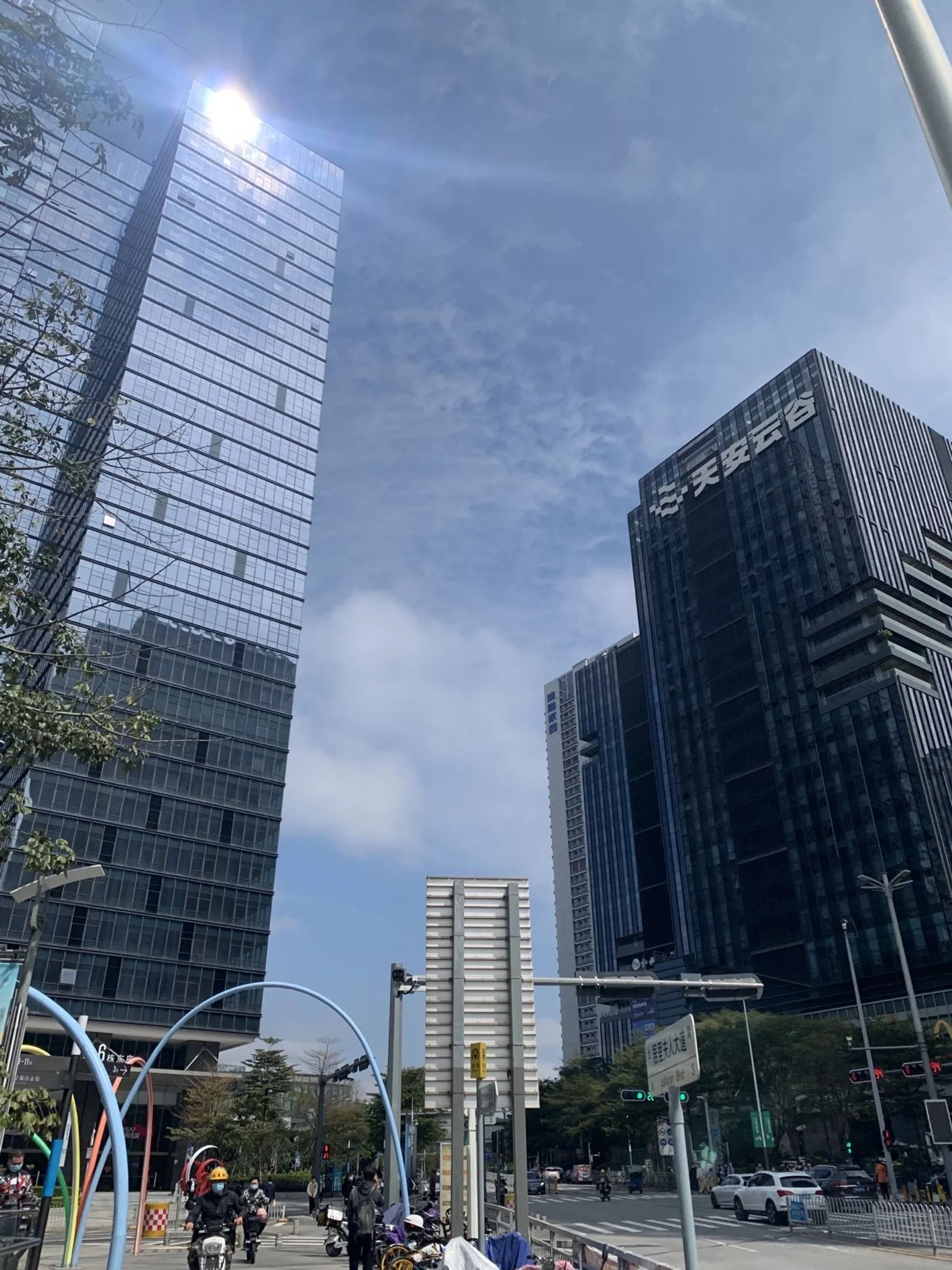 A busy city street with tall modern skyscrapers, some with glass facades, under a blue sky with scattered clouds. People are riding scooters and walking, and cars are driving on the road.