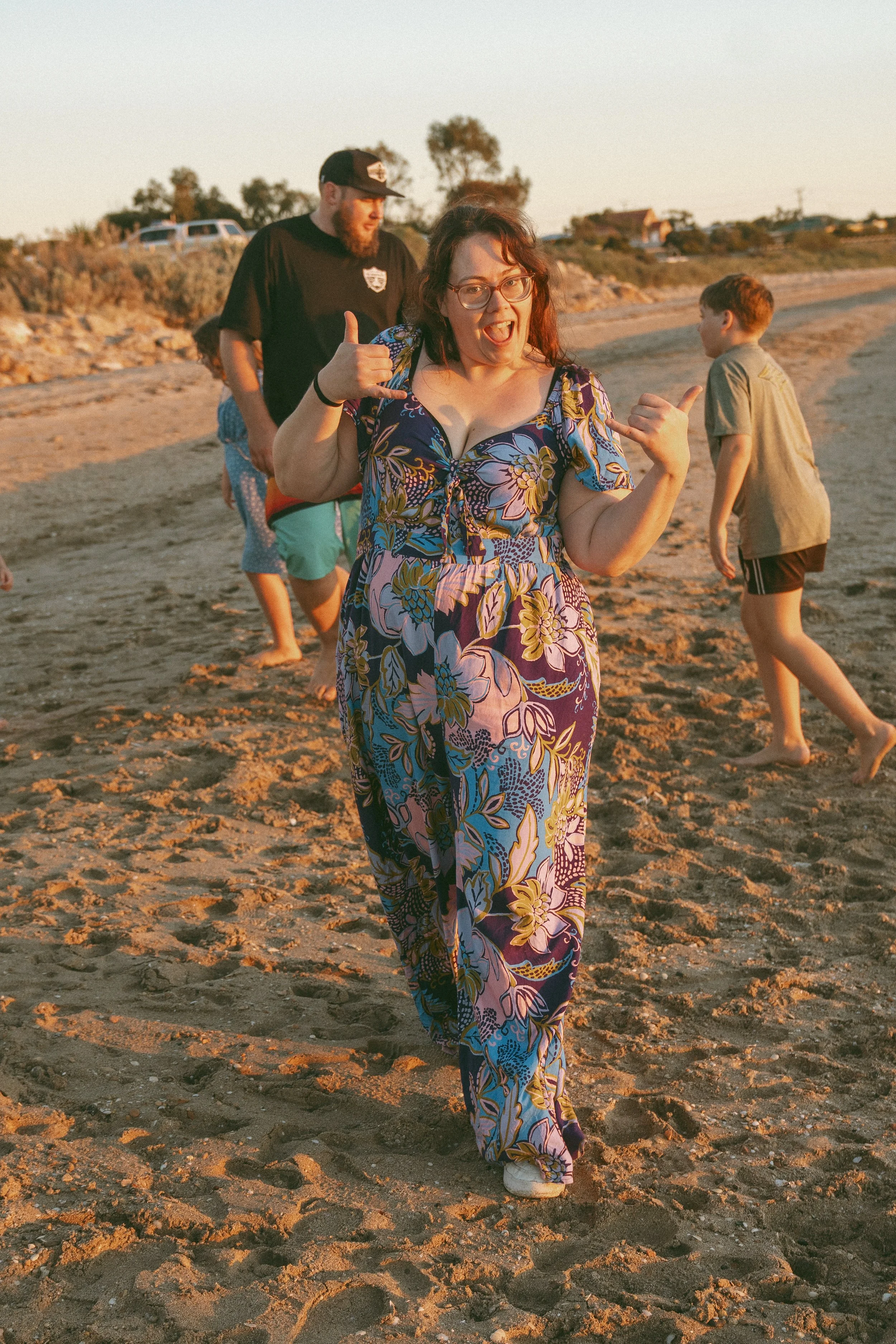 A woman in a colorful floral dress making a friendly gesture with her hand on the beach during sunset, with children and a man in casual clothing in the background.