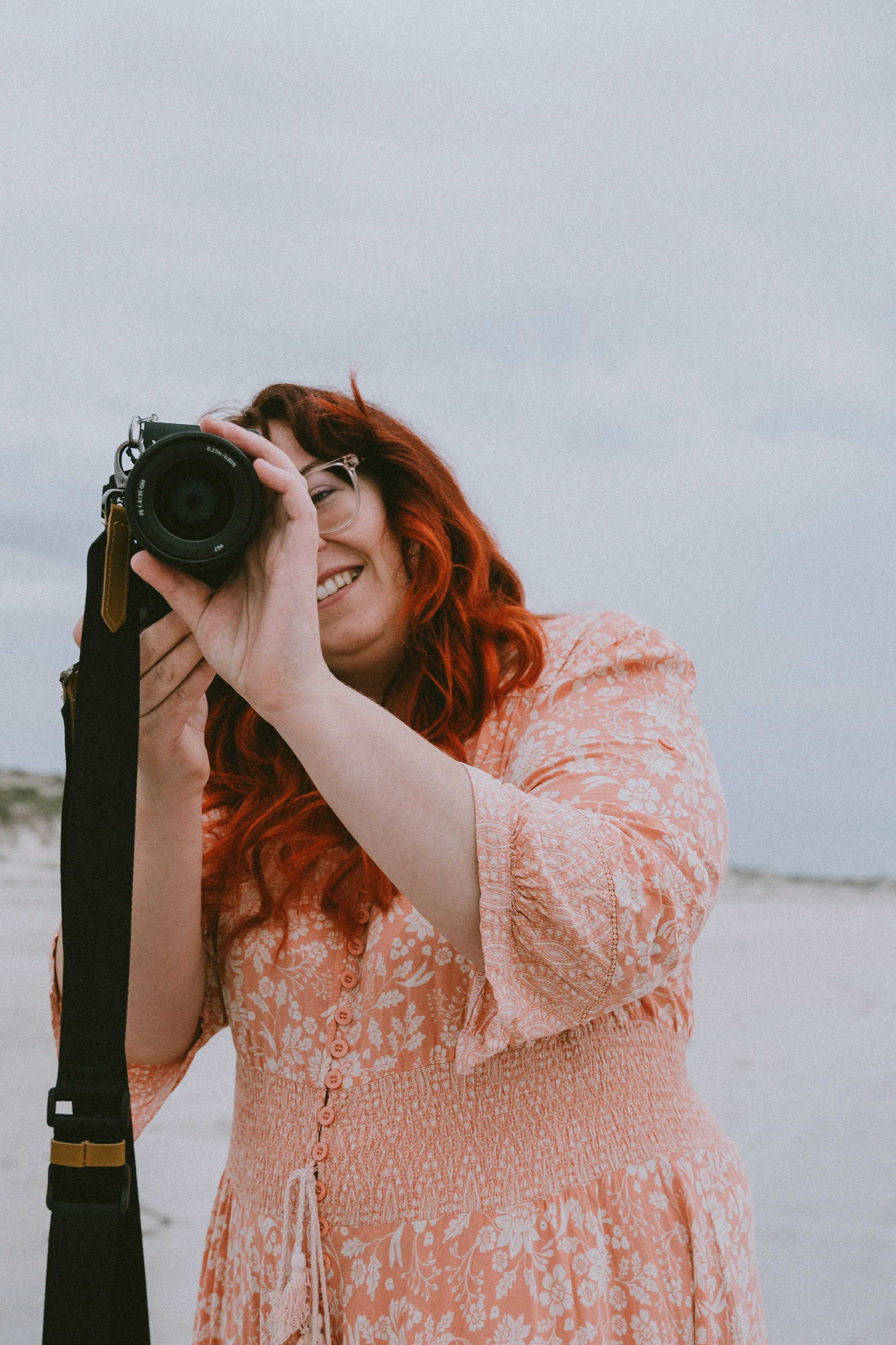 Woman with red hair taking a photo with a camera on a beach.