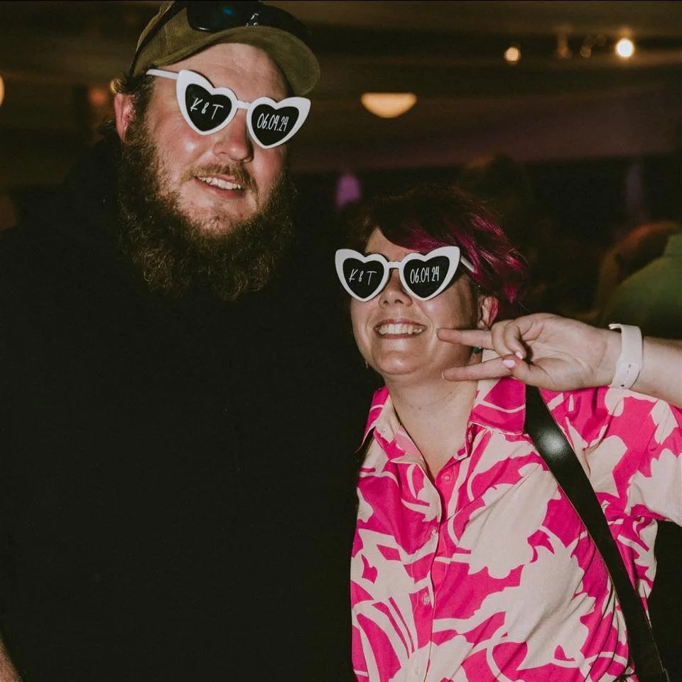 Two people wearing heart-shaped sunglasses with date and initials, smiling at a party or event. The woman is making a peace sign.