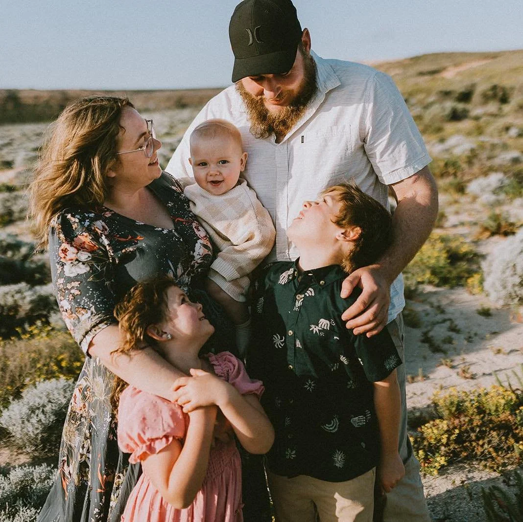 A family of six, including two children, standing outdoors on a rocky landscape with green plants, smiling and interacting with each other.