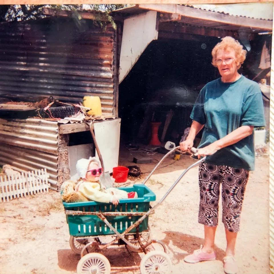 An elderly woman with curly hair and glasses pushing a toy shopping cart with a young girl sitting inside, wearing sunglasses, in front of a rustic shed.