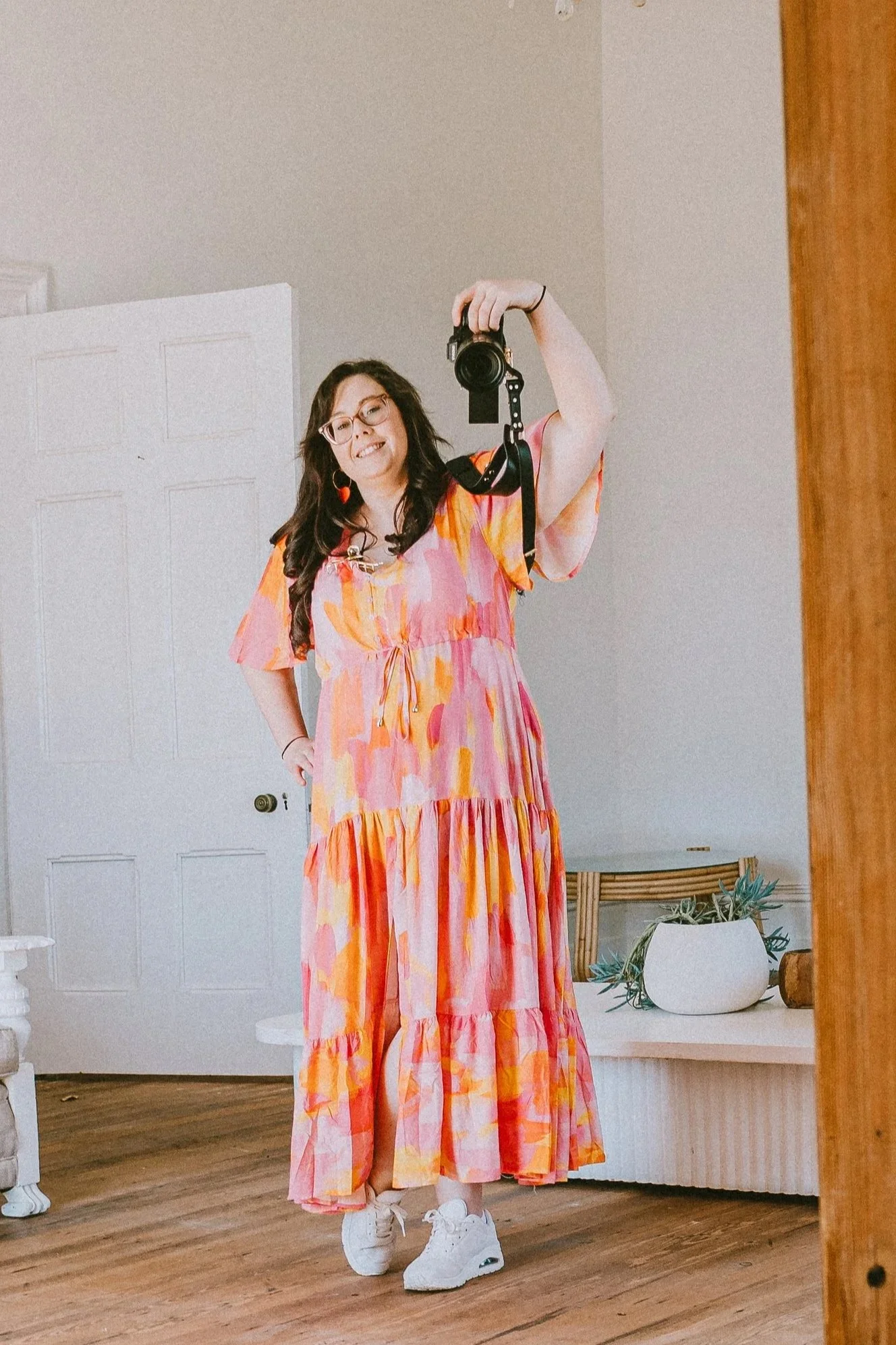 A woman in a colorful pink, orange, and yellow dress takes a mirror selfie inside a room. She has long dark hair, wears glasses and earrings, and stands on a wooden floor.