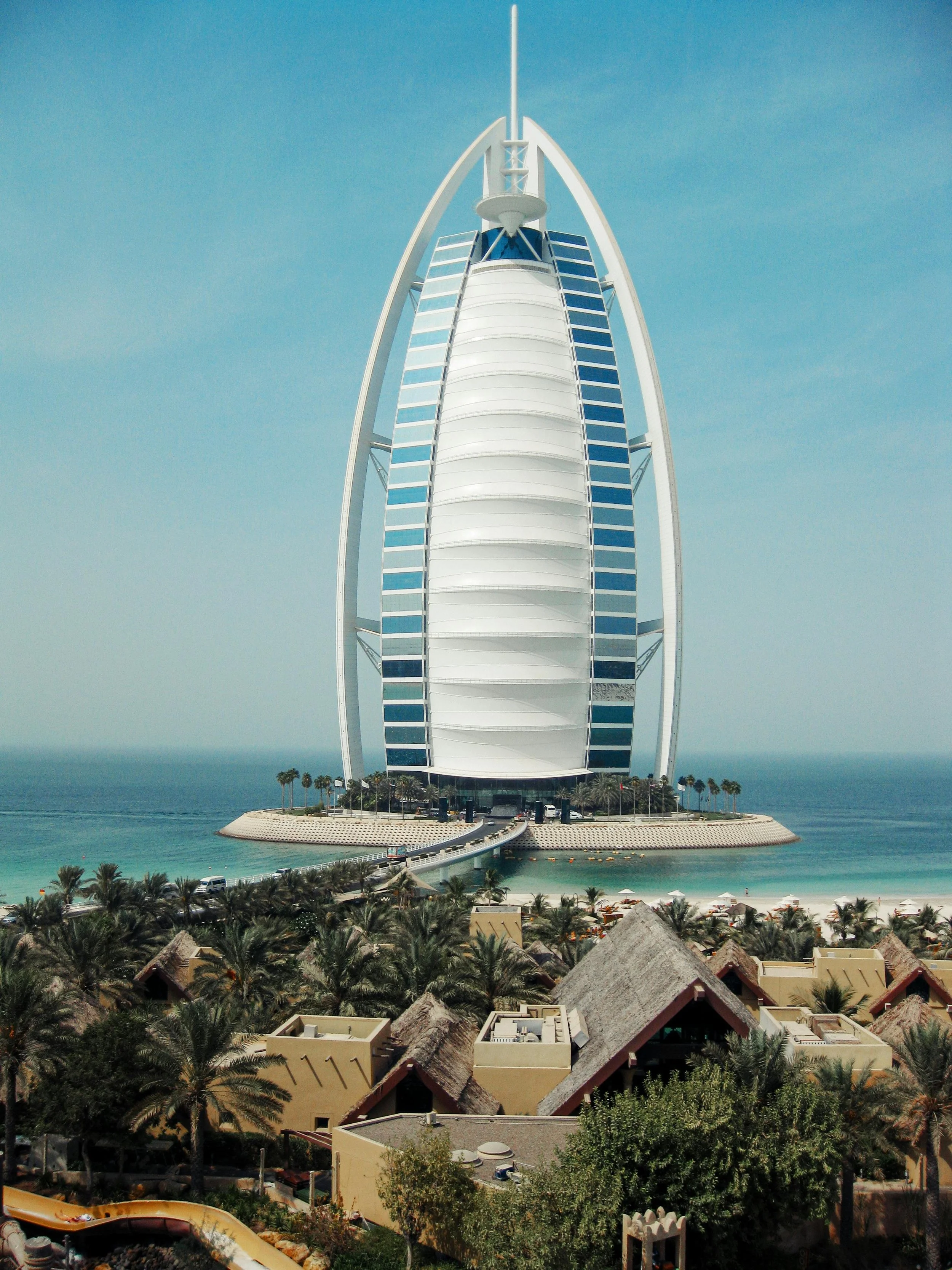 View of the Burj Al Arab hotel in Dubai, a sail-shaped luxury hotel on the Arabian Gulf, with palm trees and thatched rooftops in the foreground.