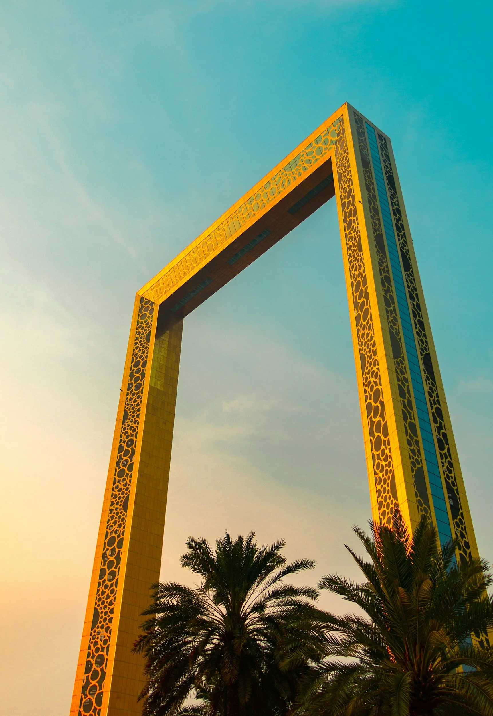 The Dubai Frame, a large rectangular gold-colored structure with a patterned border, stands against a blue sky with palm trees in the foreground.
