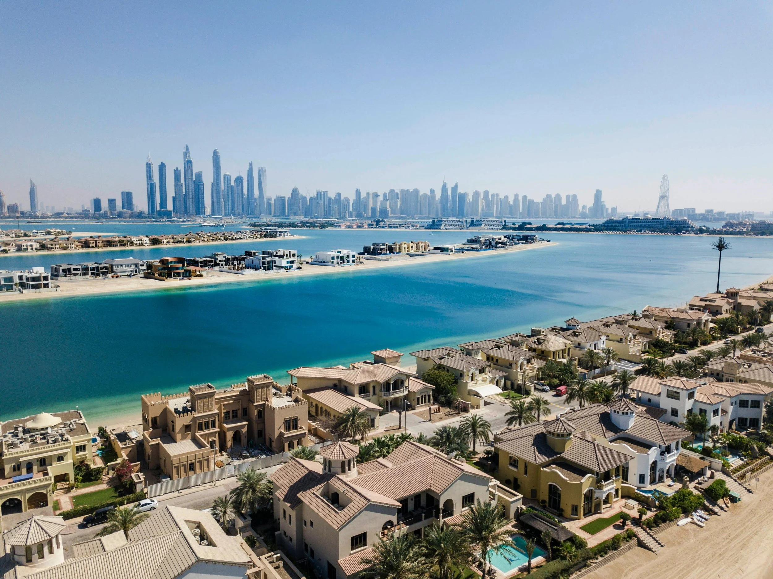 Aerial view of Dubai skyline with boats on water and residential houses with pools in the foreground.