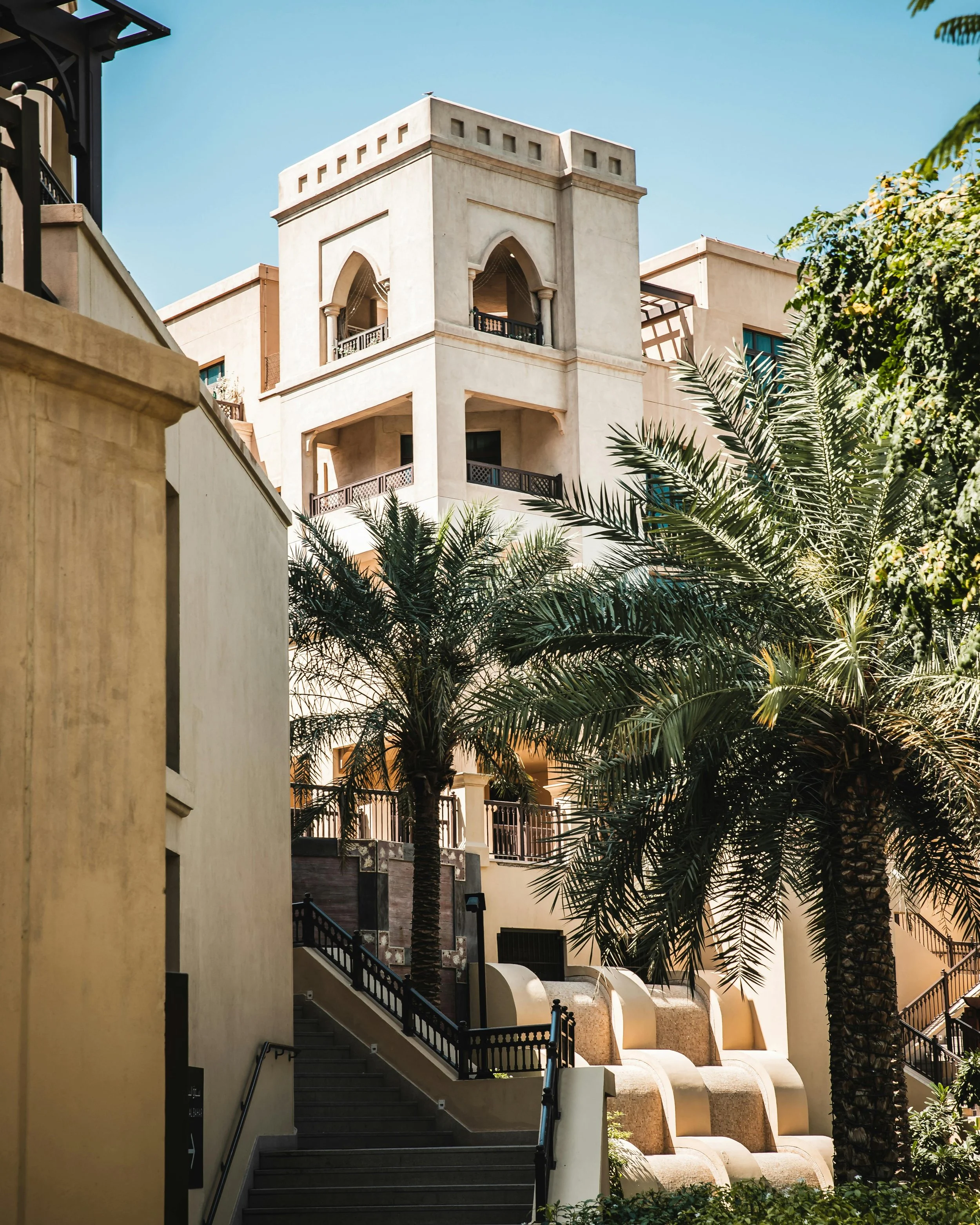 Multiple beige and cream-colored buildings with balconies and arches, surrounded by green palm trees, under a clear blue sky.