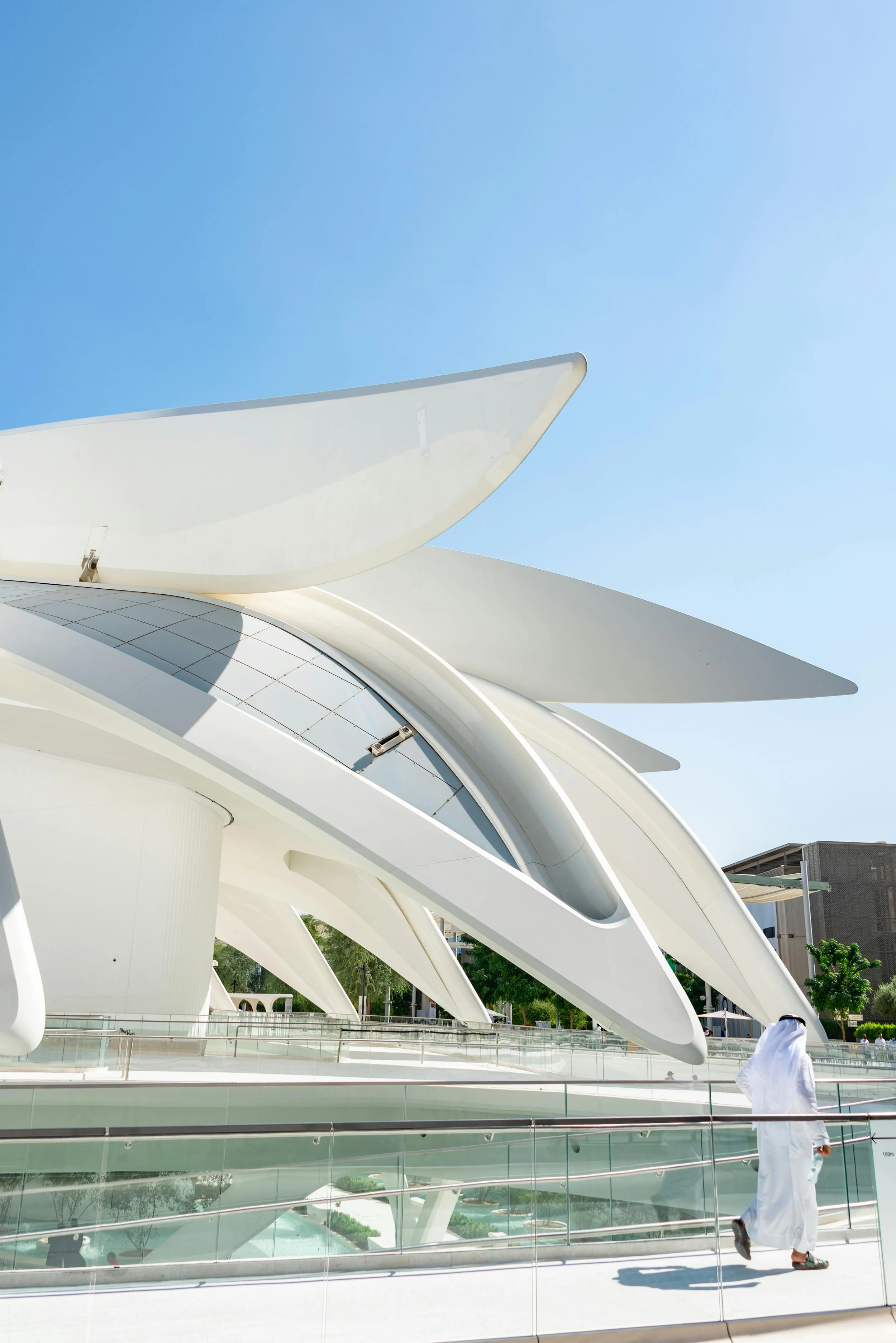 Modern futuristic building with white curved, wing-like structures and glass roof panels, with a person in white traditional Middle Eastern attire walking nearby under a clear blue sky.