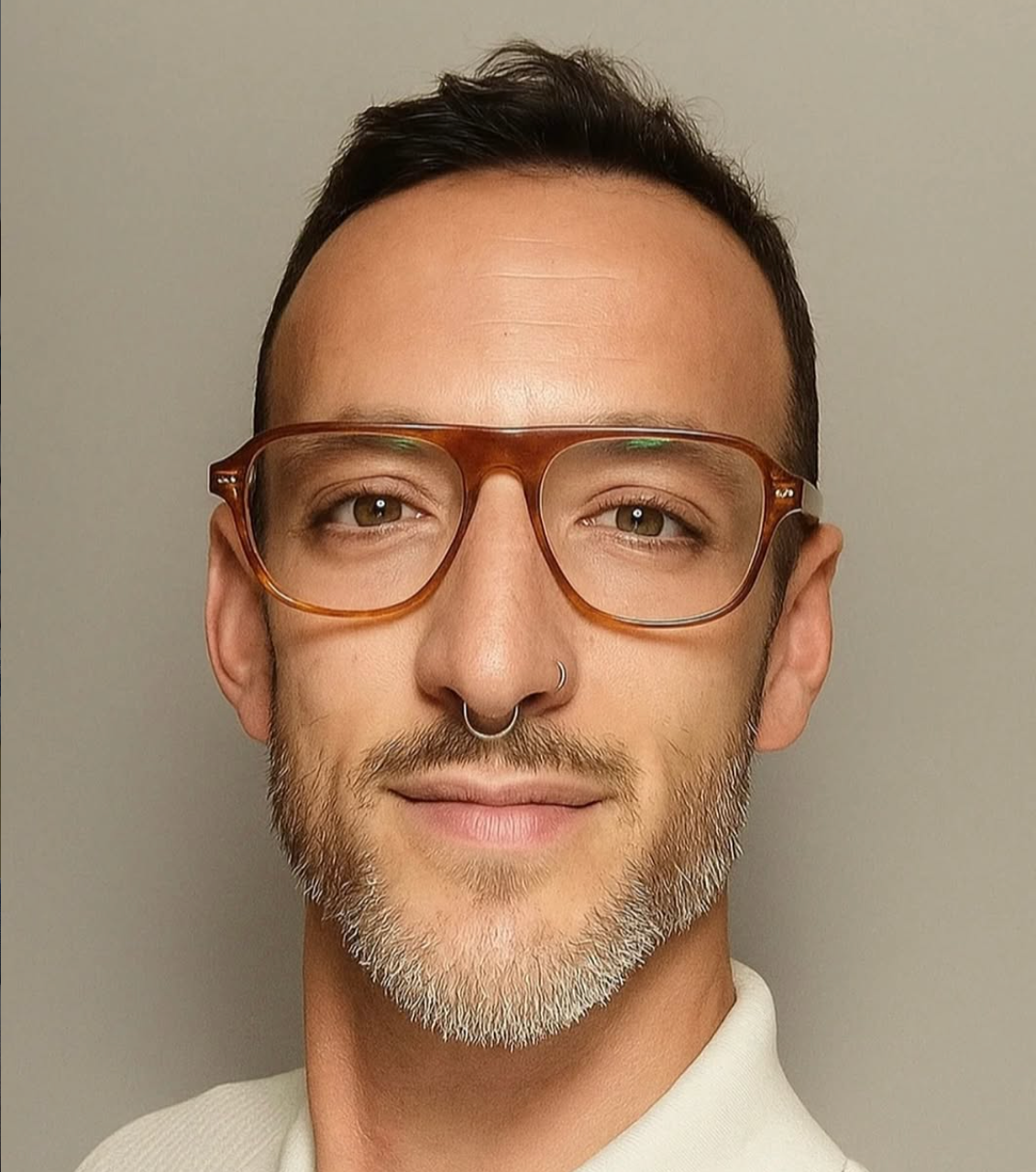 Retrato de un hombre con gafas color carey, barba gris, septum piercing y sonrisa ligera, contra fondo gris.