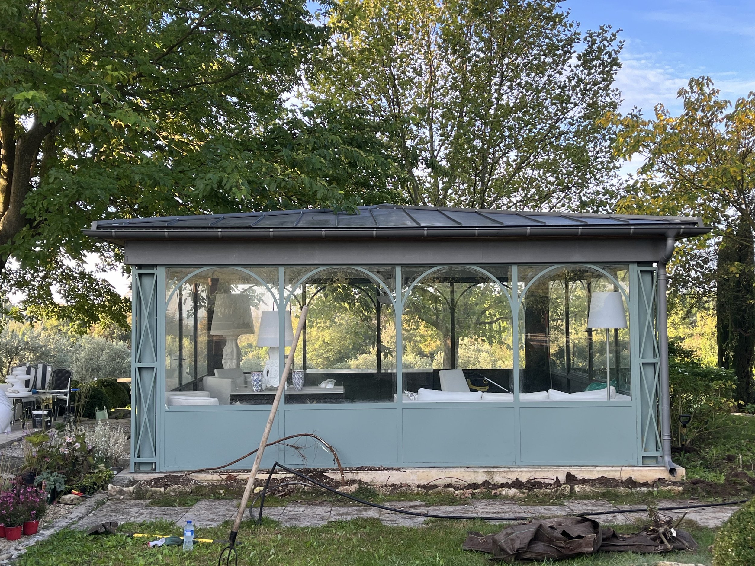 A screened porch or garden room with glass panels, light blue frame, and a black metal roof, surrounded by trees and greenery.