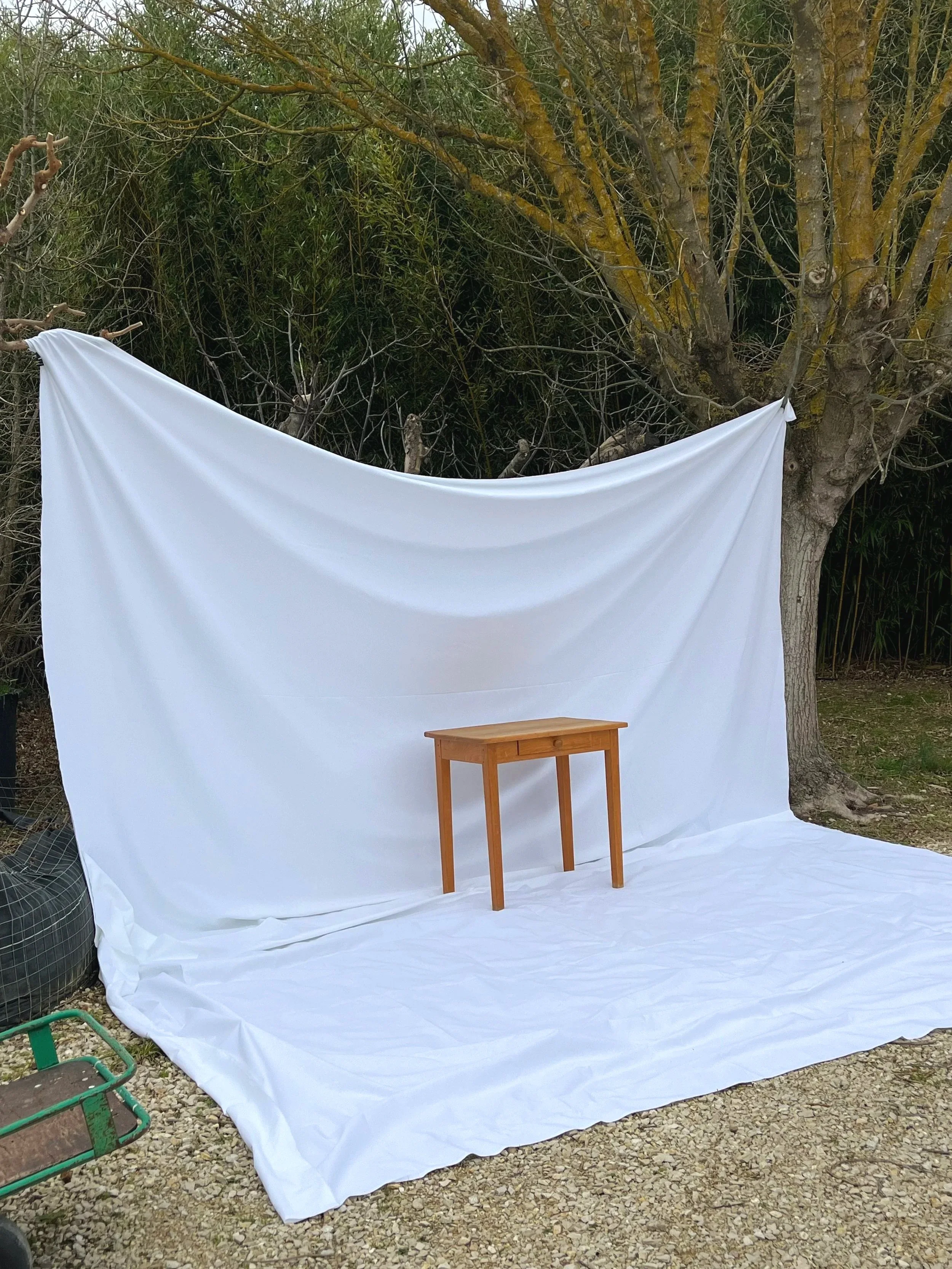 Outdoor scene with a large white fabric backdrop attached to a tree, with a small wooden table in front of it on a gravel ground, and a green wheelbarrow to the left.