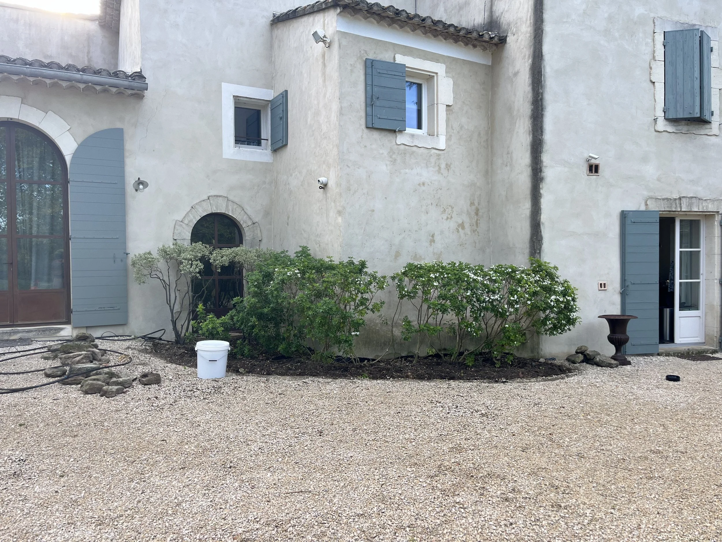 Exterior of a European-style house with white stucco walls, gray shutters, and arched windows, surrounded by gravel and greenery under a clear sky.