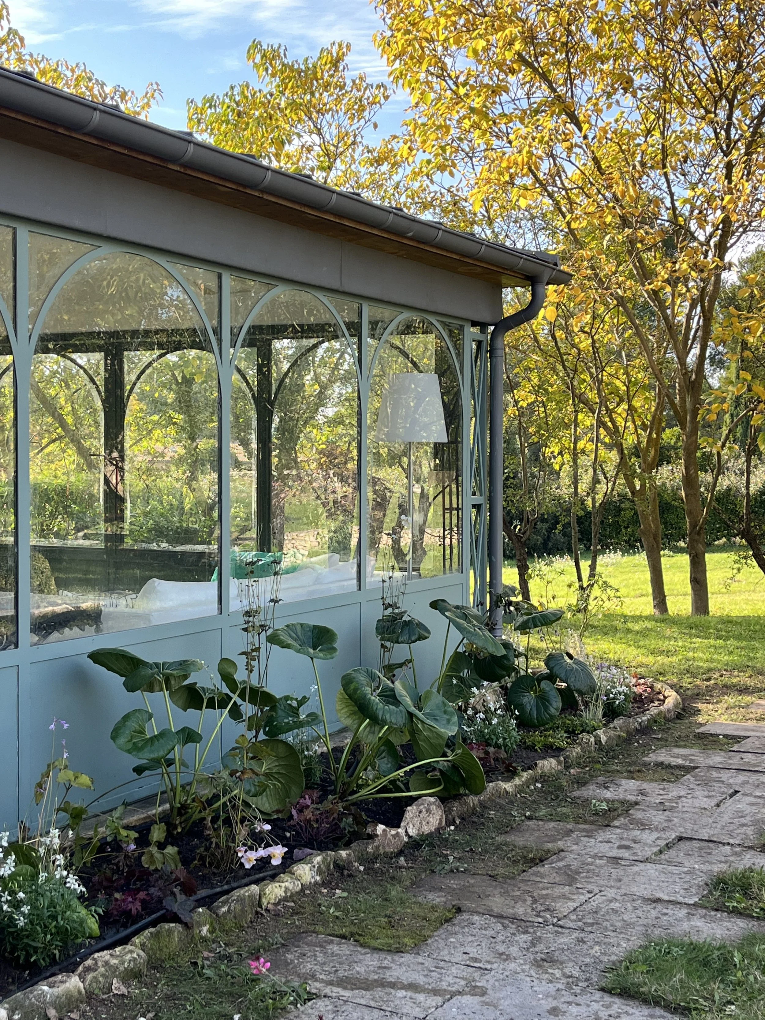A garden greenhouse with a glass exterior, surrounded by plants and flowers, with trees and a grassy yard in the background on a sunny day.
