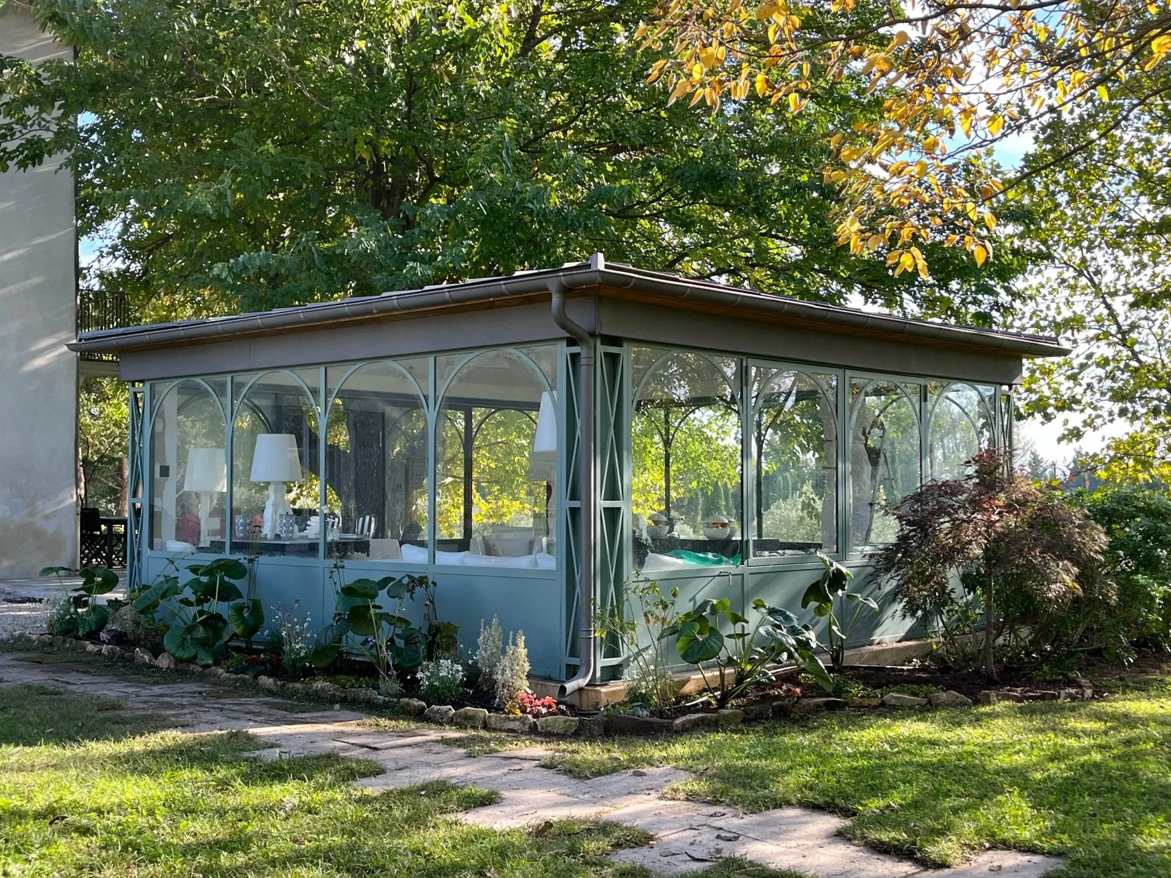 A glass greenhouse with arched windows in a garden, surrounded by plants and a stone pathway.