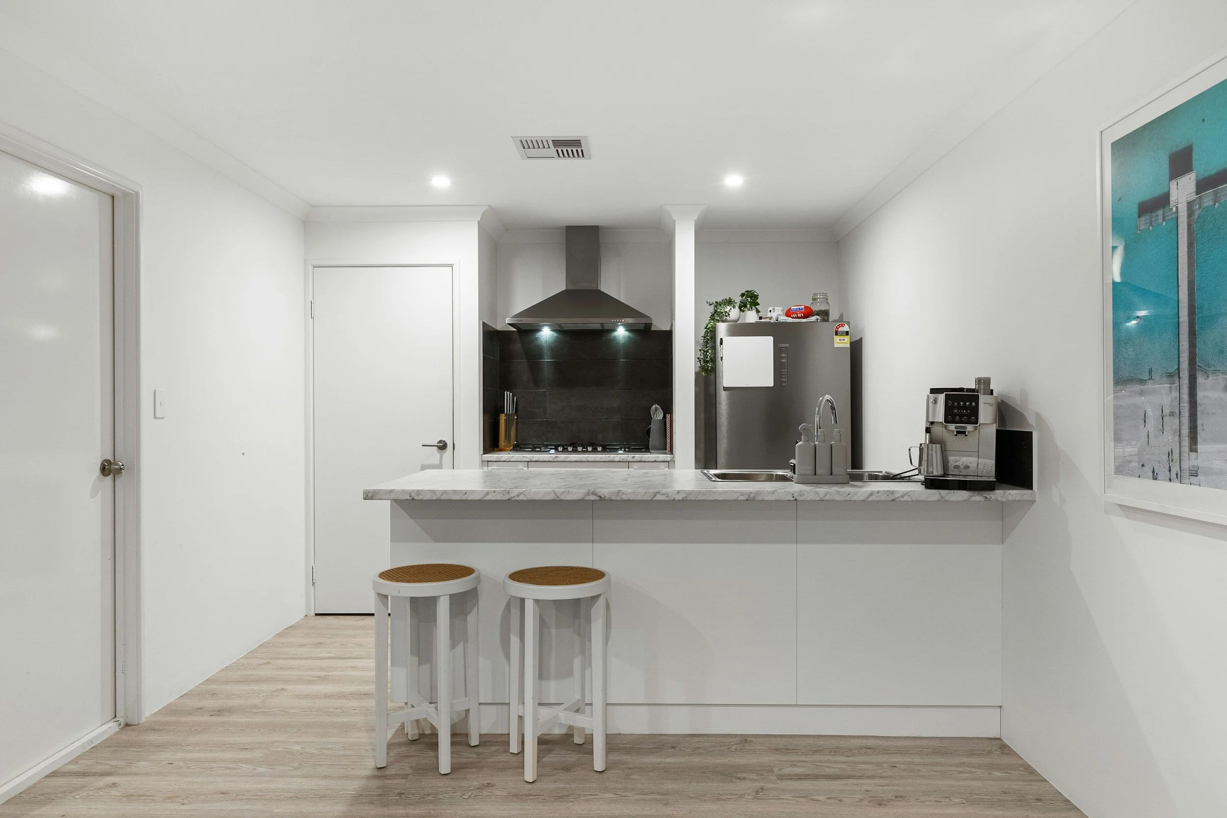 Modern kitchen with white walls and cabinetry, marble countertop, stainless steel appliances including a refrigerator, coffee machine, and stove, with two round bar stools in front.