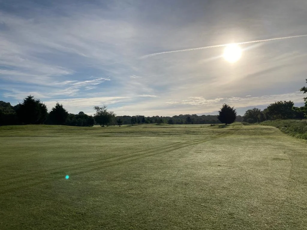 Halfpenny Green golf course under a bright sun with a partly cloudy sky and several trees scattered along the fairway.