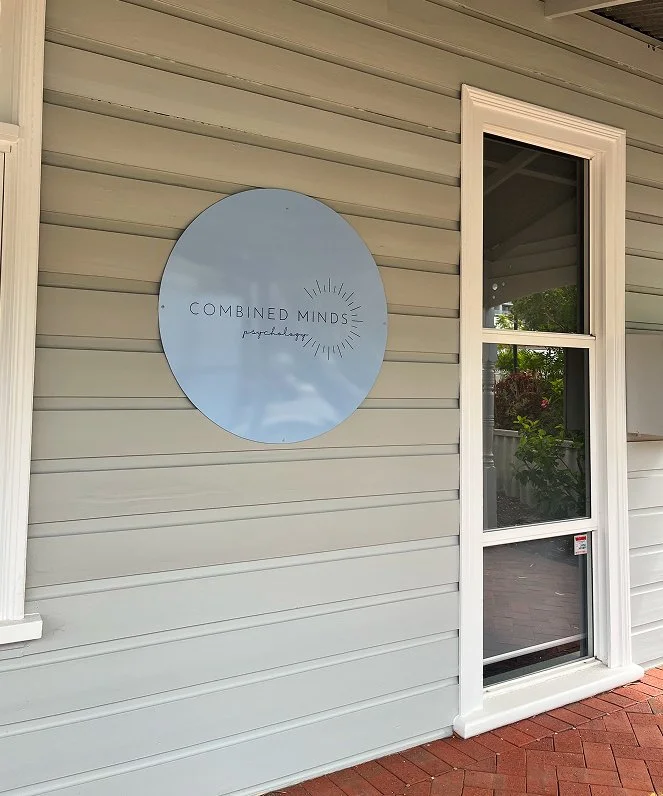 Exterior view of a building with beige siding, a white-framed door with a screen, and a round sign that reads "Combined Minds psychotherapy" with sunburst design.