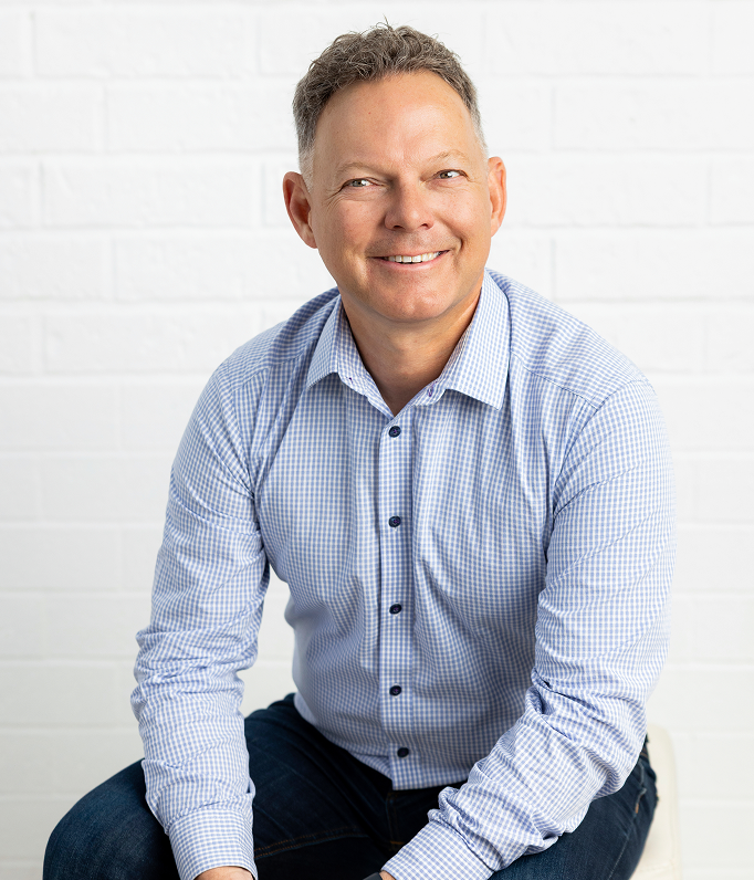 A smiling middle-aged man with short gray hair, wearing a light blue checkered shirt, sitting against a white brick wall background.