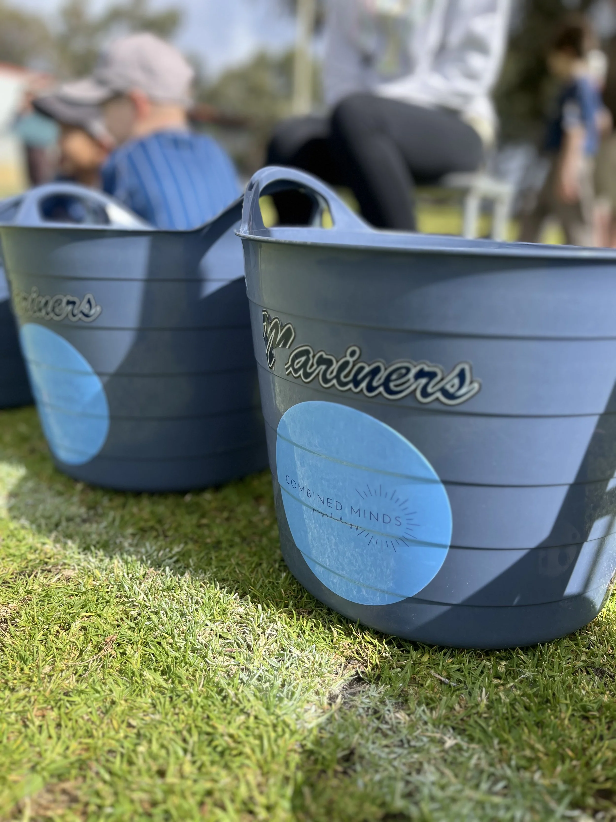 Close-up of two blue buckets labeled 'Mariners' sitting on grass, with blurred people in the background at an outdoor event.