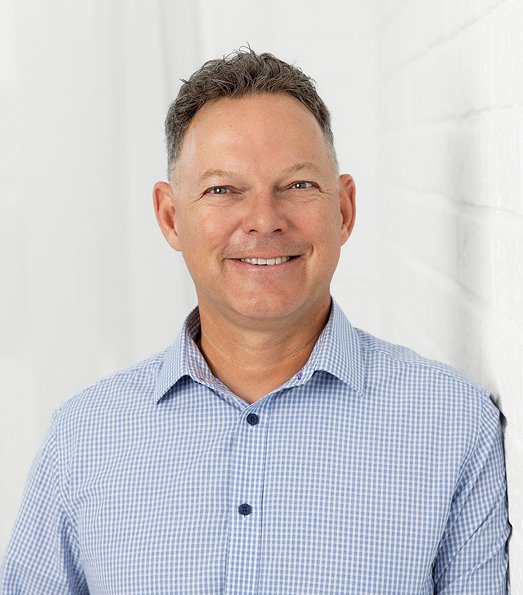 A smiling middle-aged man with short, dark hair, wearing a light blue checkered button-up shirt, standing against a white wall.