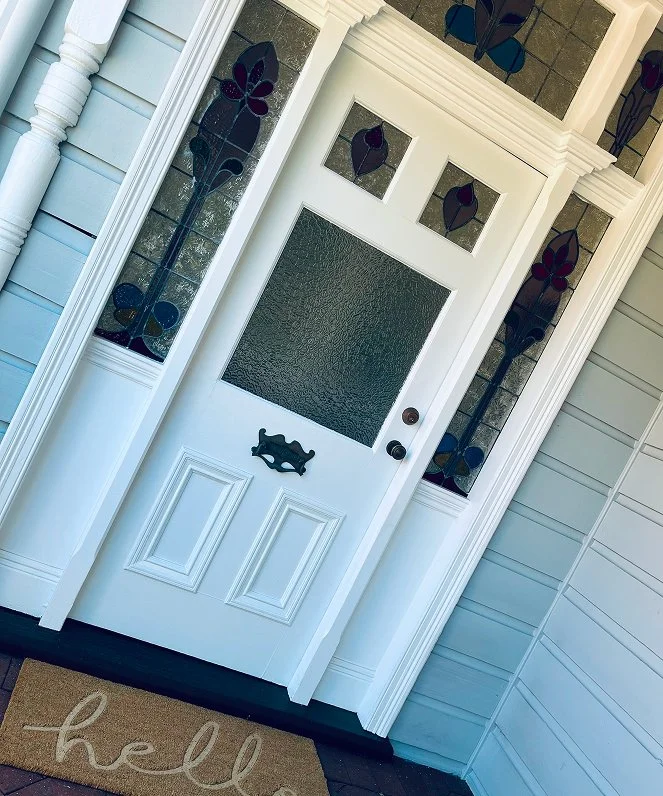Front porch with white door featuring a frosted glass window and black handles, decorative stained glass side panels with floral designs, and a light brown doormat with 'hello' written in cursive.
