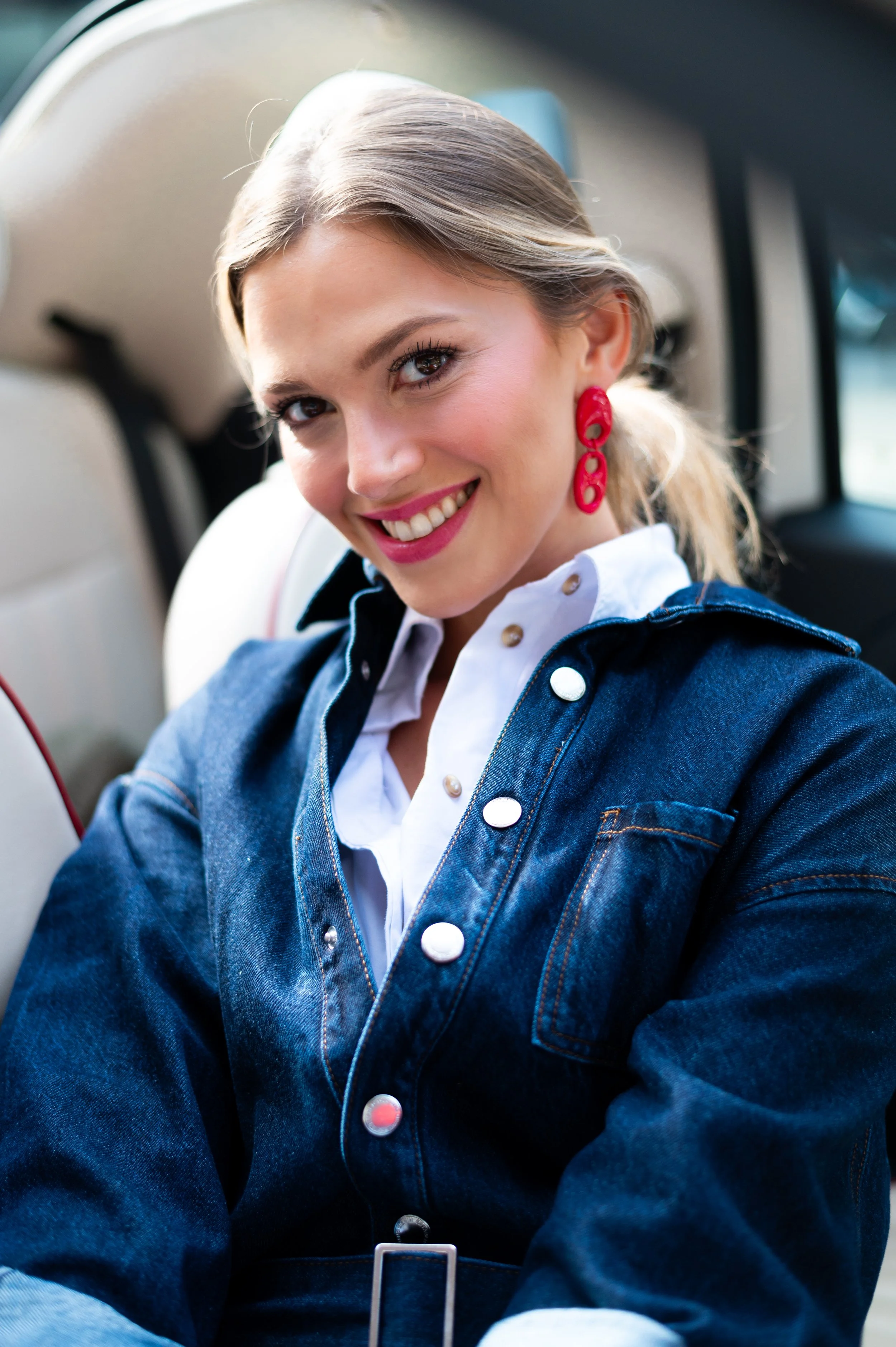 Une jeune femme souriante, portant des boucles d'oreilles rouges et une chemise blanche sous une veste en jean, assise à l'intérieur d'une voiture.