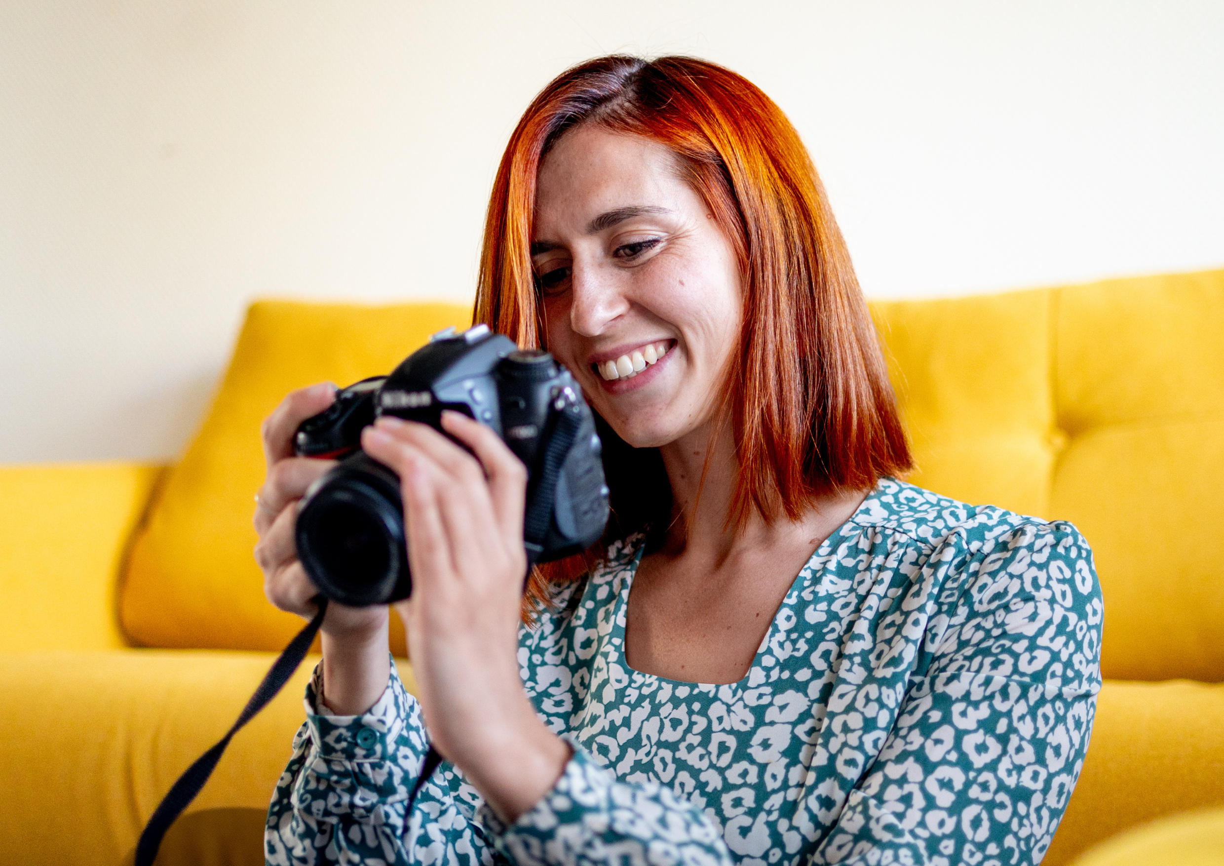 Une femme aux cheveux roux, souriante, regarde un appareil photo numérique qu'elle tient dans ses mains, assise sur un canapé jaune.