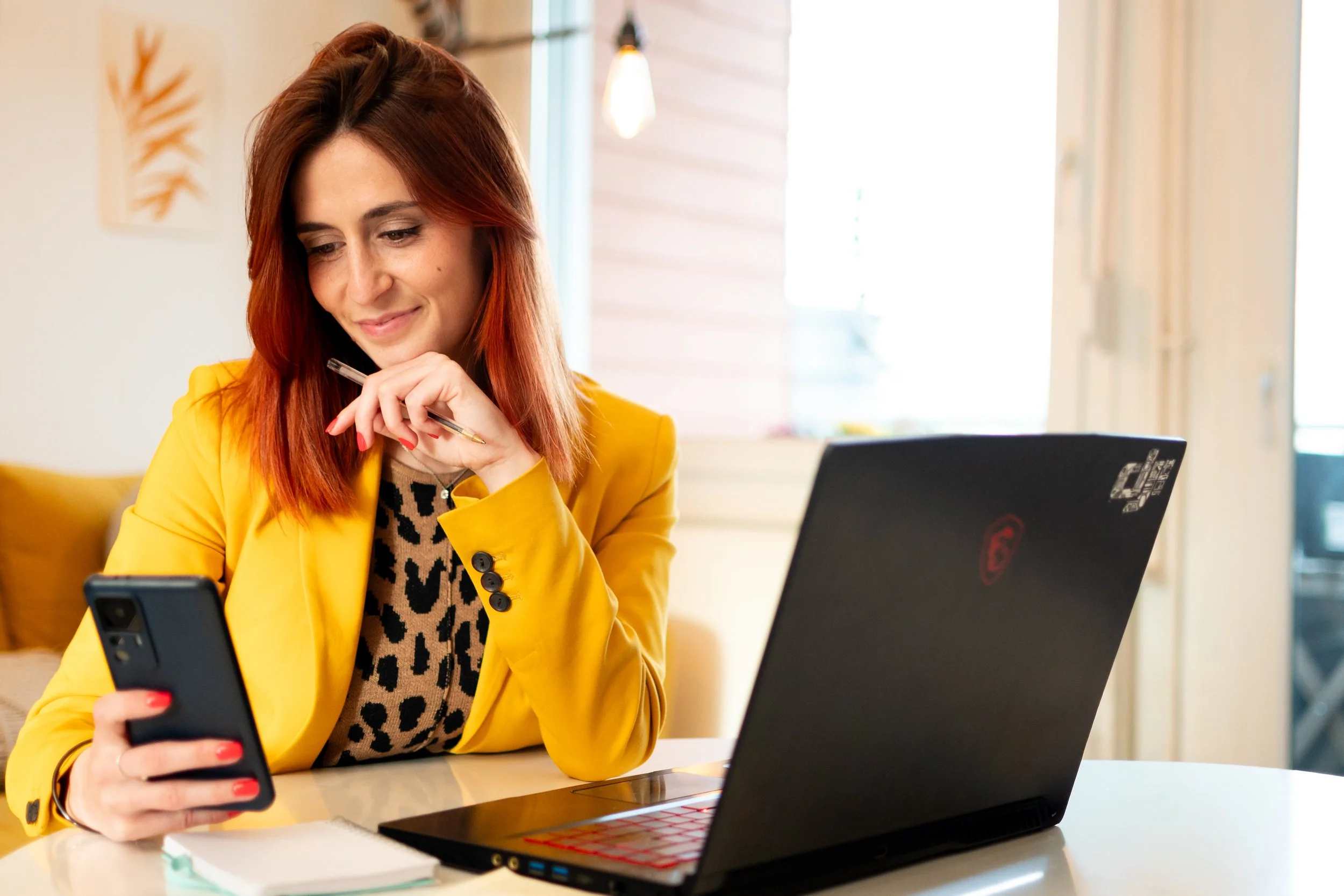 Femme souriante avec cheveux roux portant une veste jaune, utilisant un smartphone dans un espace intérieur bien éclairé avec un ordinateur portable ouvert sur une table.