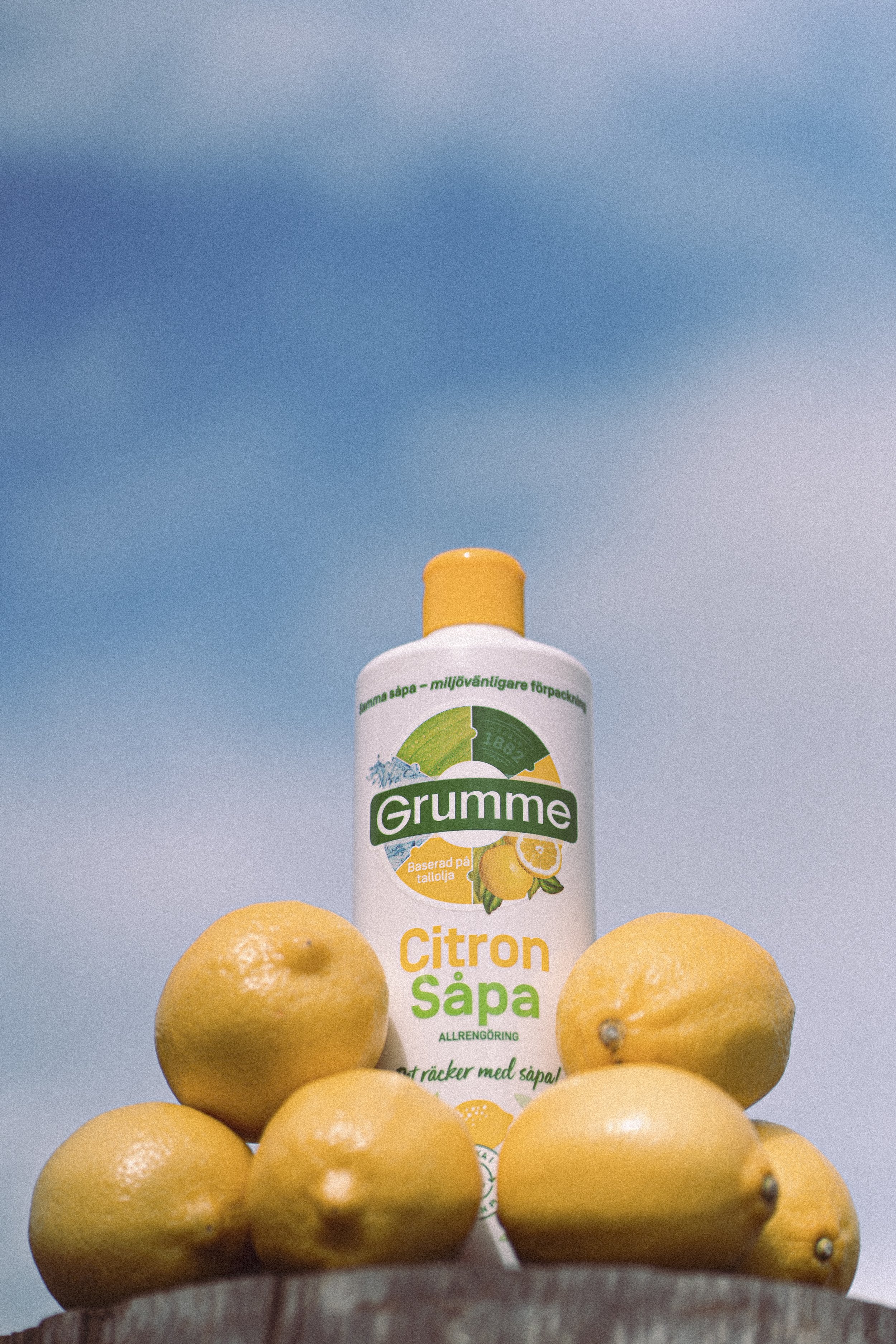 A bottle of Grumme lemon soap surrounded by whole lemons against a blue sky background.