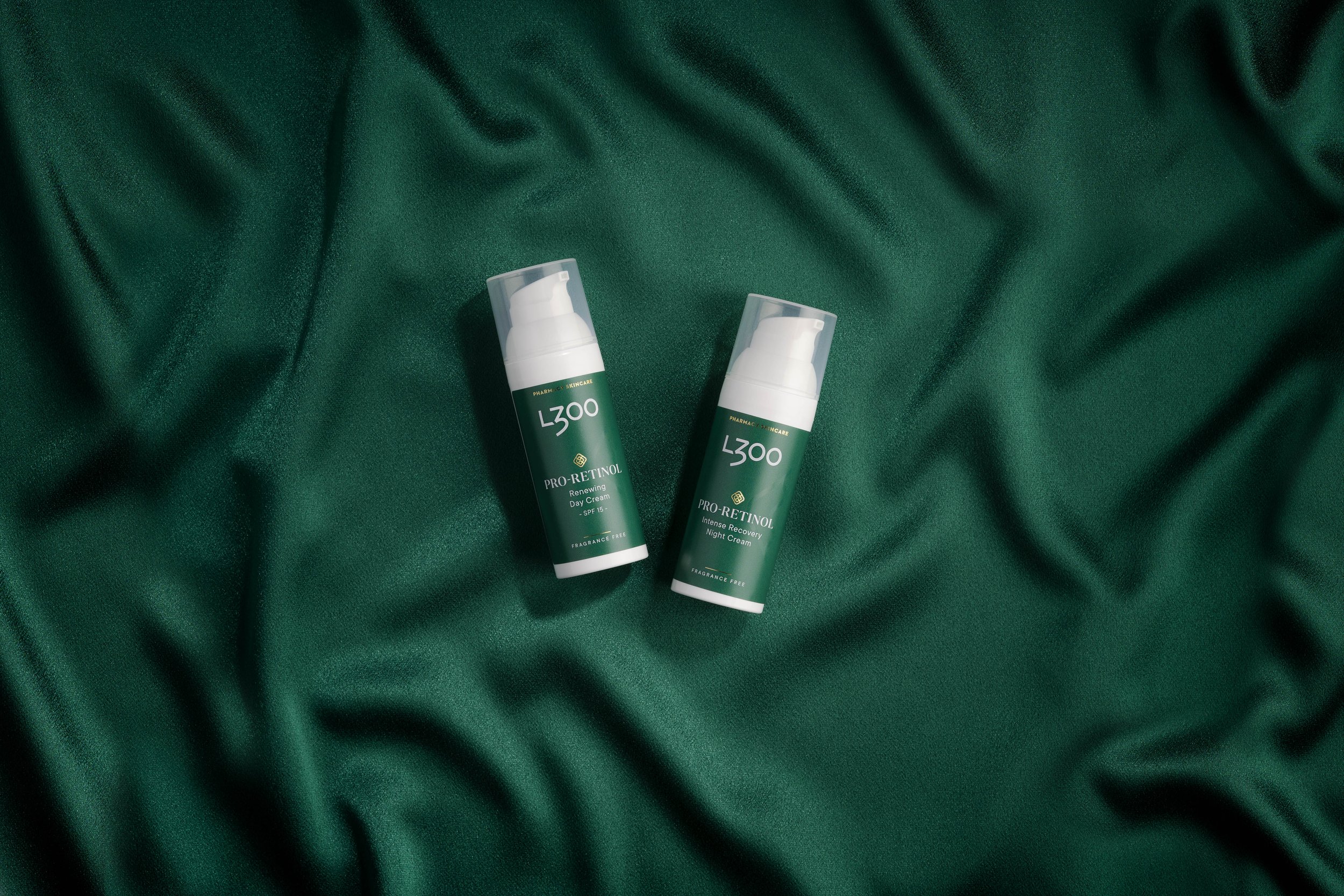 Two green bottles of L300 skincare products placed on green fabric, one labeled 'Pro-Retinol Renewing Day Cream' and the other 'Pro-Retinol Intense Recovery Night Cream'.