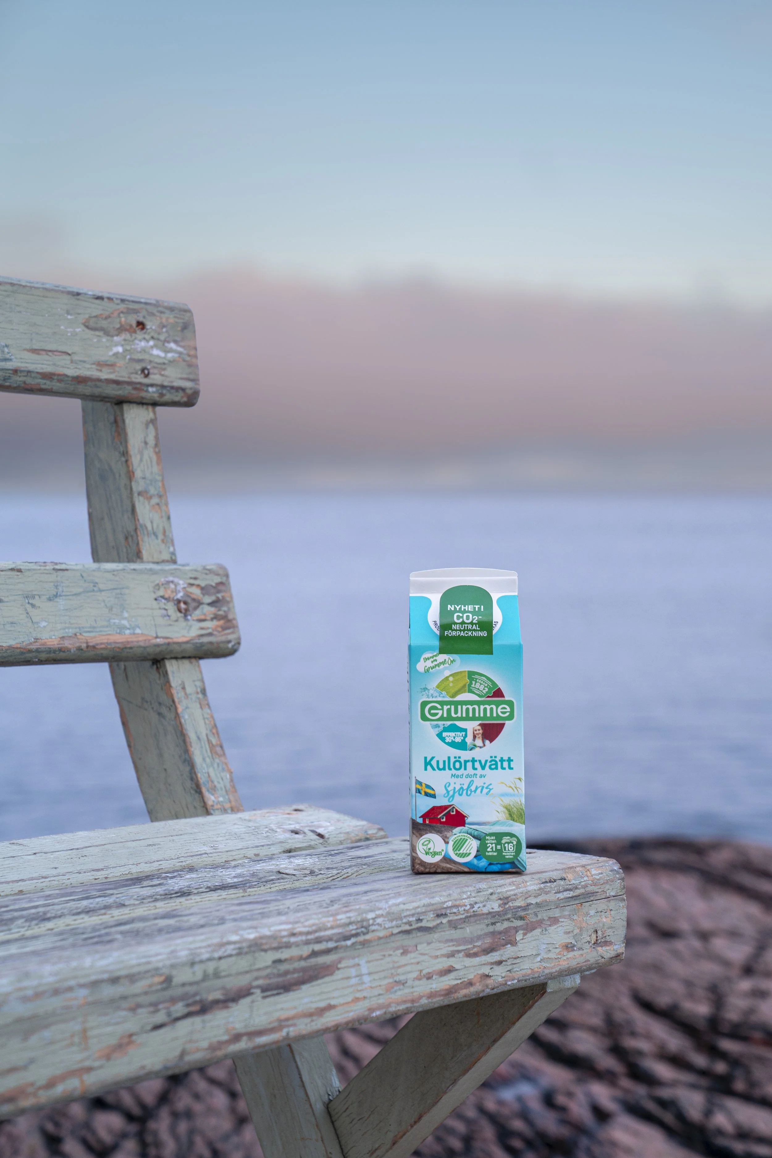 A carton of Grumme dishwashing liquid on a weathered wooden bench by the water with rocks, overlooking a calm body of water and a pink and blue sky at sunset.