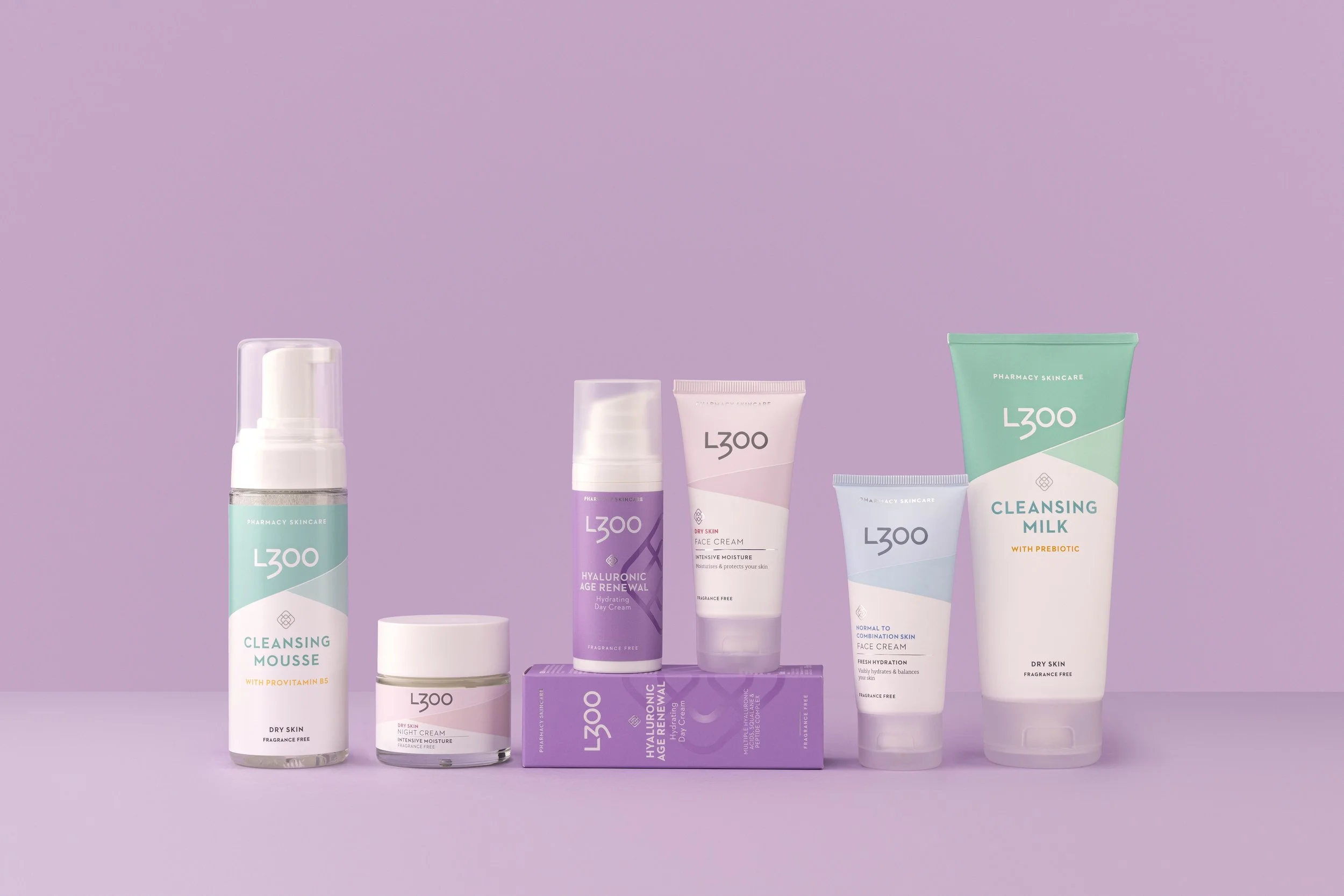 Line of skincare products including cleansing mousse, night cream, hyaluronic acid serum, face cream, another face cream, and cleansing milk against a pastel purple background.