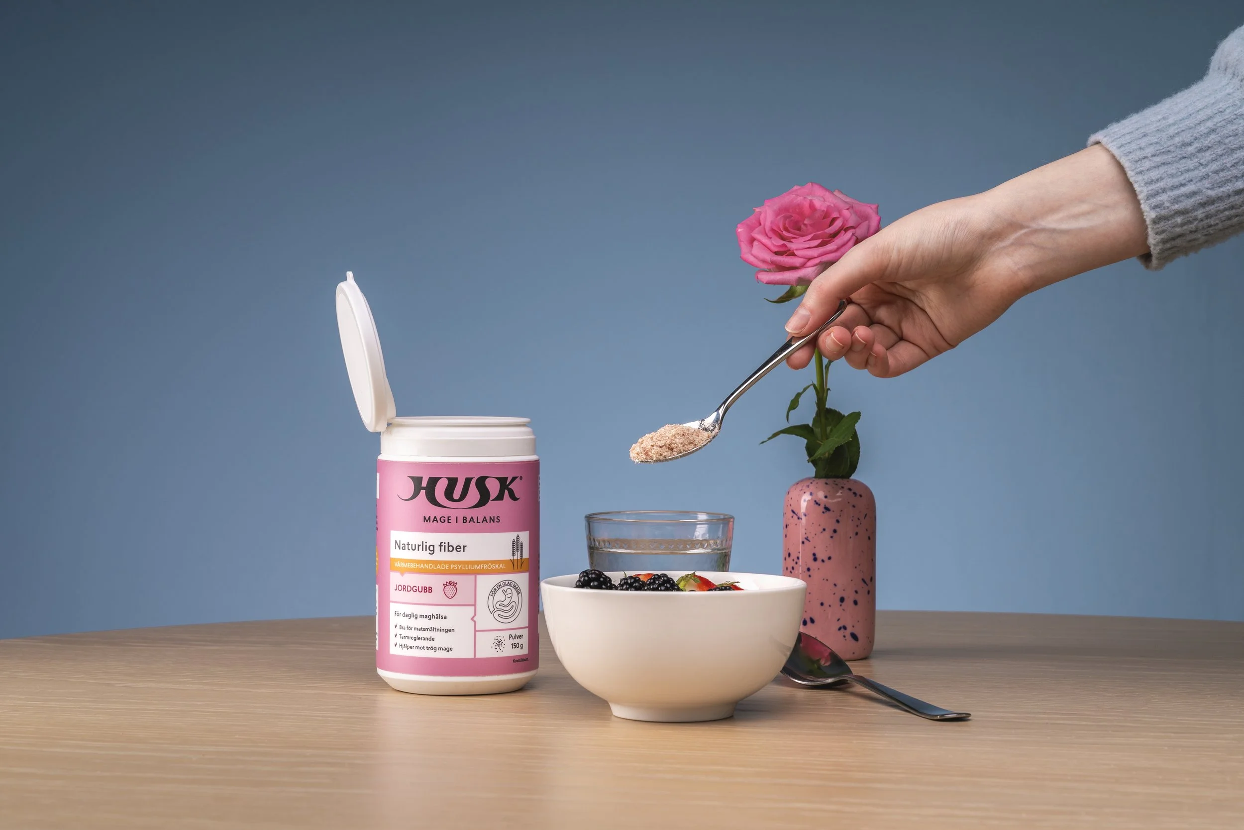 A person with a gray sleeve sprinkling powder into a bowl of yogurt with fruit, on a wooden table, with a pink flower in a vase, a glass of water, and a container labeled 'Husk' in the background.