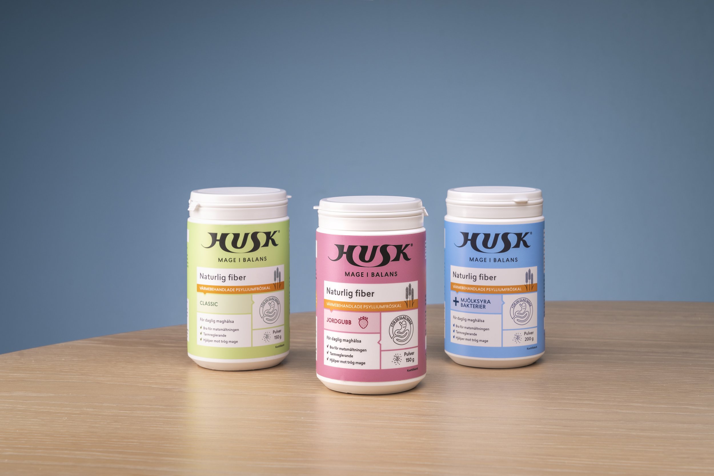 Three containers of HUSK fiber supplements on a wooden table against a blue background, with one in green labeled 'Classic', one in pink labeled 'Jordgubb', and one in blue labeled 'Mjölksyra Bakterier'.