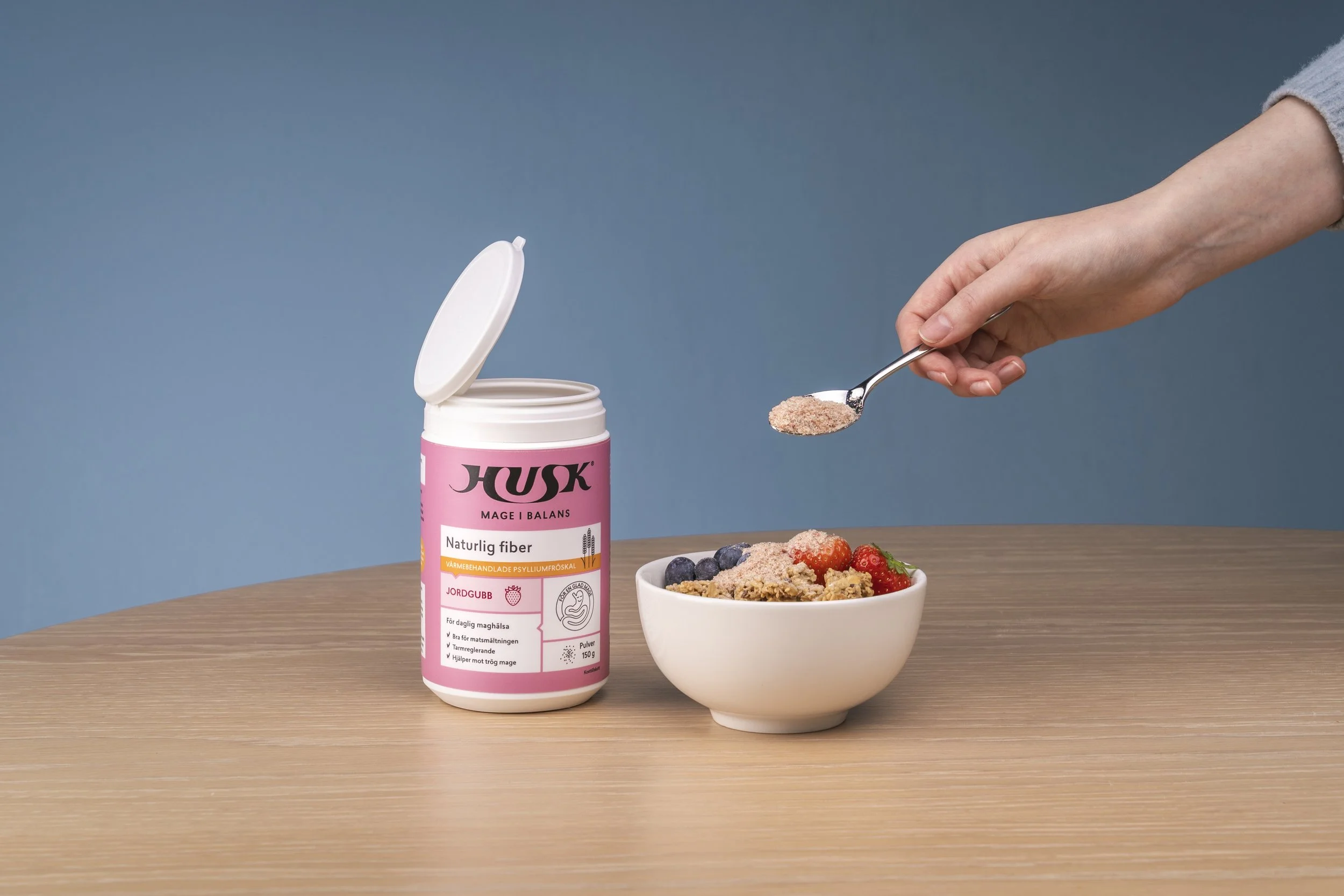 A person's hand sprinkling powder from a spoon over a bowl of cereal with berries, next to a pink container labeled 'Husk' on a wooden table with a blue background.