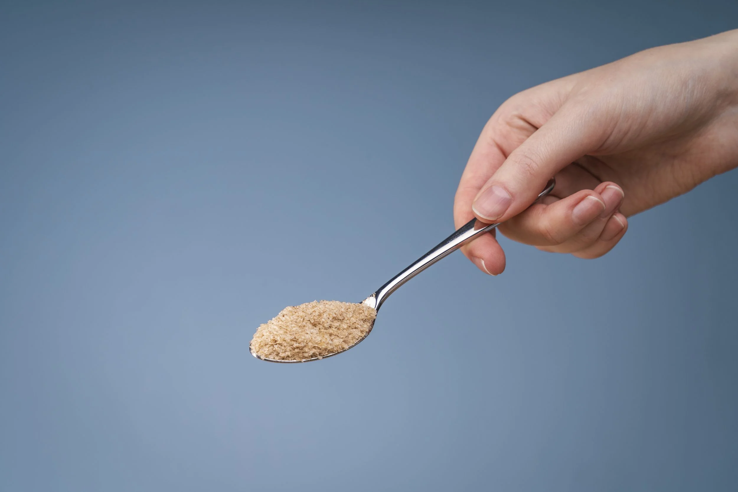 Hand holding a spoon with brown powder against a blue background.