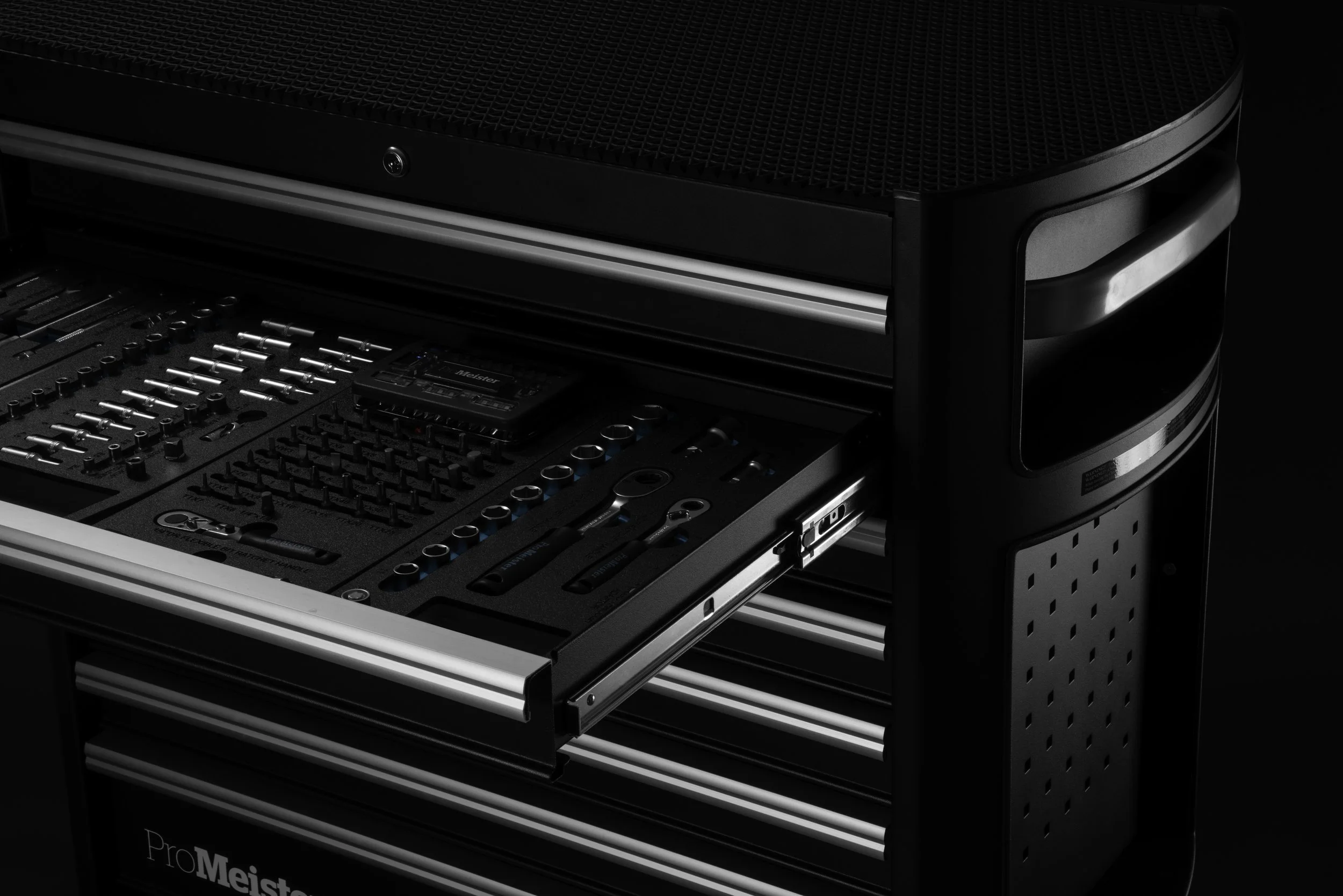 A black professional tool storage cart with multiple drawers. The top drawer is open, revealing a sound and light control panel, tools, and audio equipment.