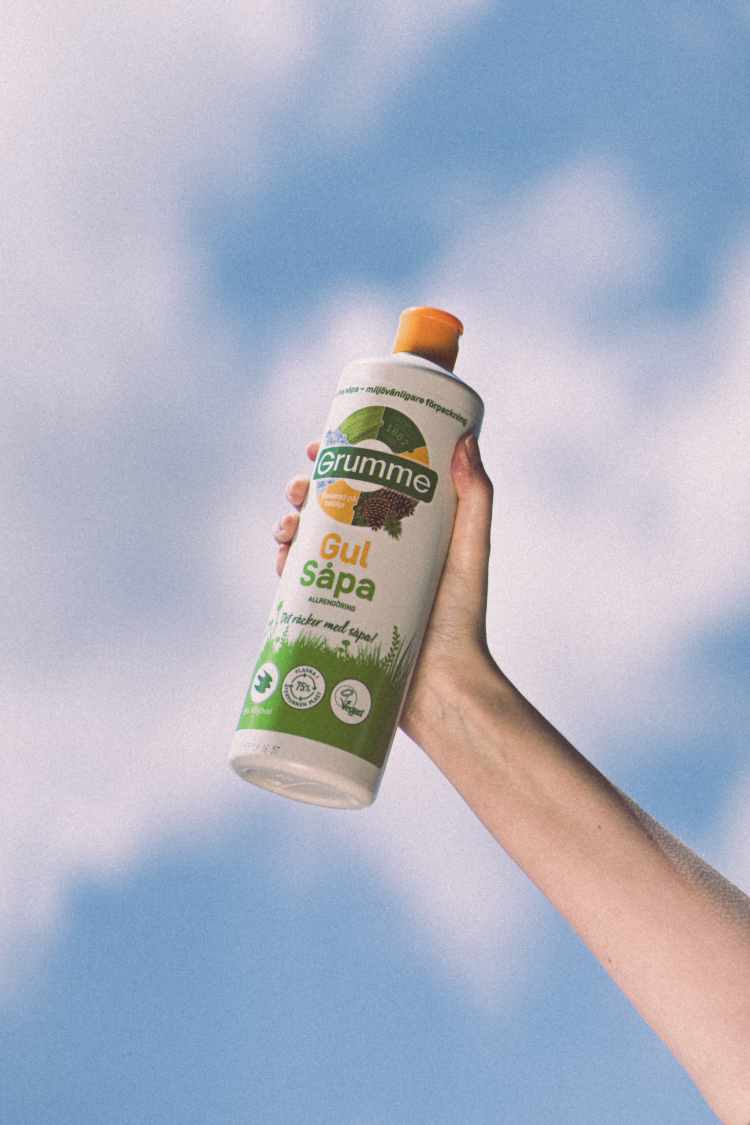 A hand holding a bottle of Grumme Gul Såpa, an all-purpose cleaning product, outdoors against a partly cloudy sky.