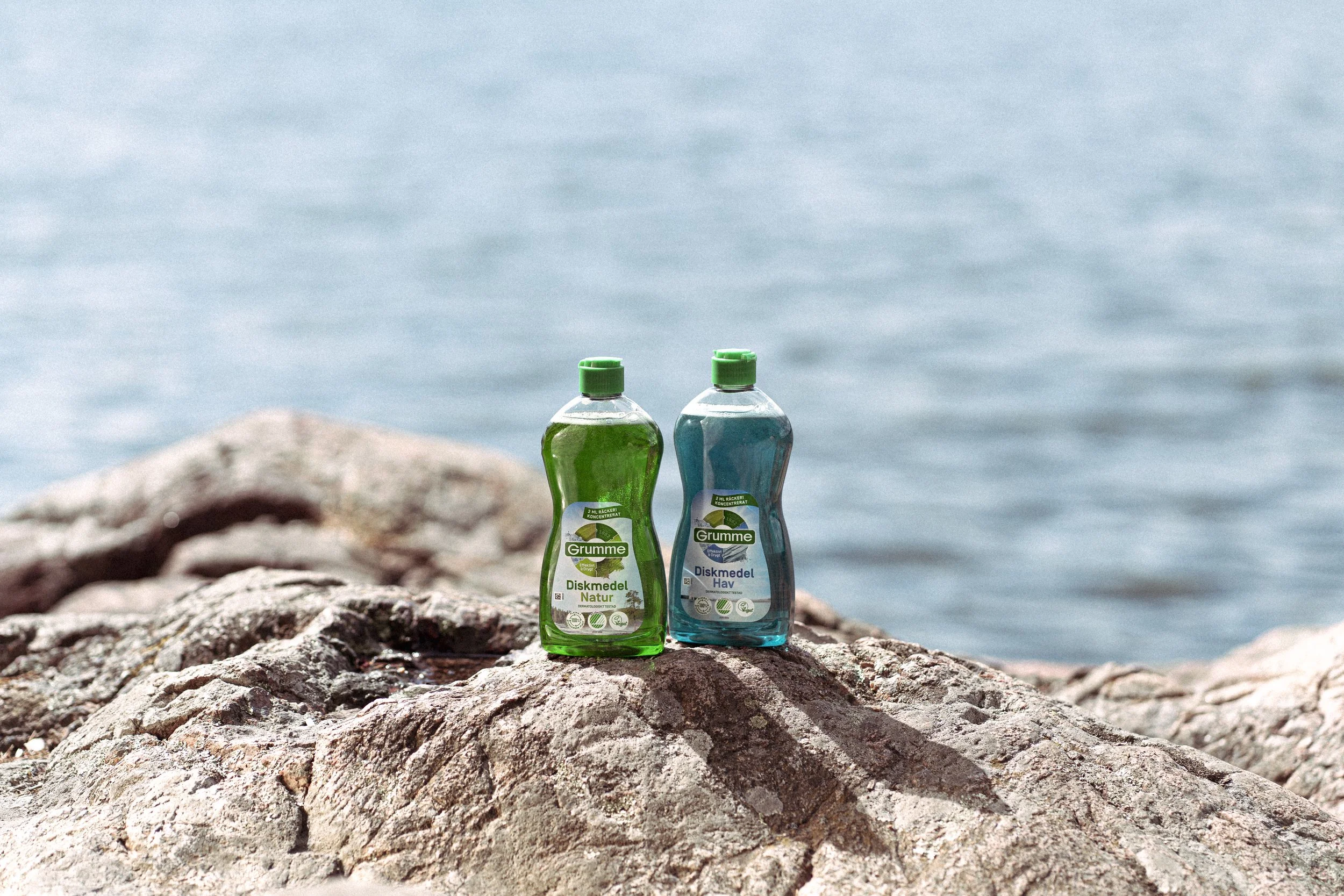 Two bottles of cleaning product, one green and one blue, placed on a rocky surface near the water with a blurred body of water in the background.