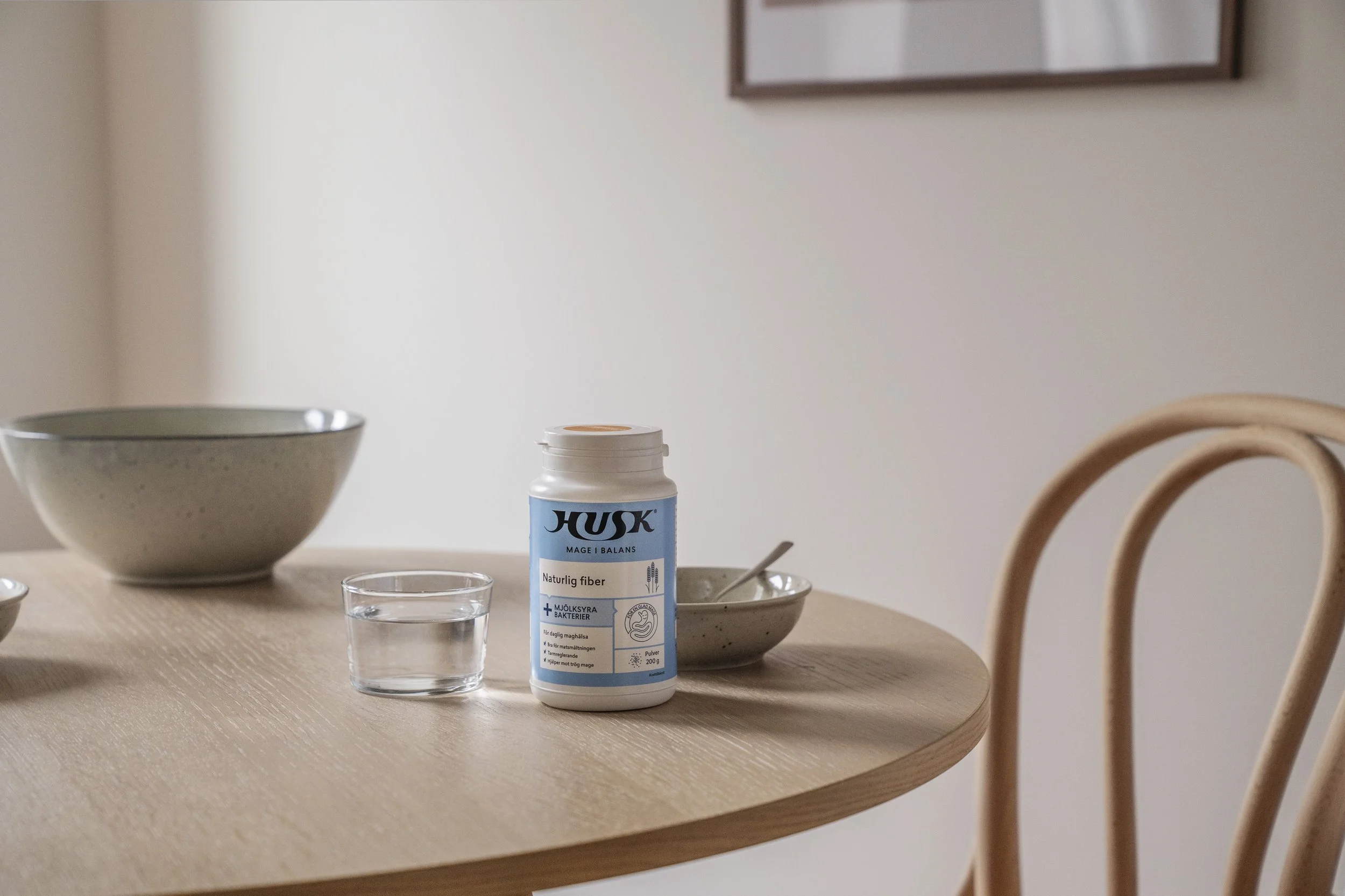 A wooden dining table with a white container of dietary fiber, a glass of water, and two bowls, one larger and one smaller, in a minimalist kitchen or dining area with a light-colored wall and a framed picture hanging on it.