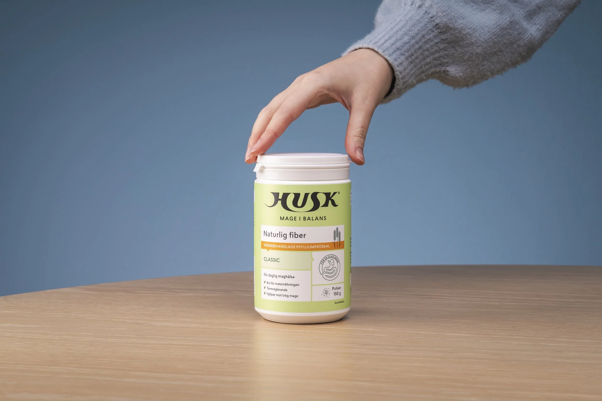 A hand in a gray sweater reaching for a container of HUSK natural fiber dietary supplement on a wooden table against a blue background.